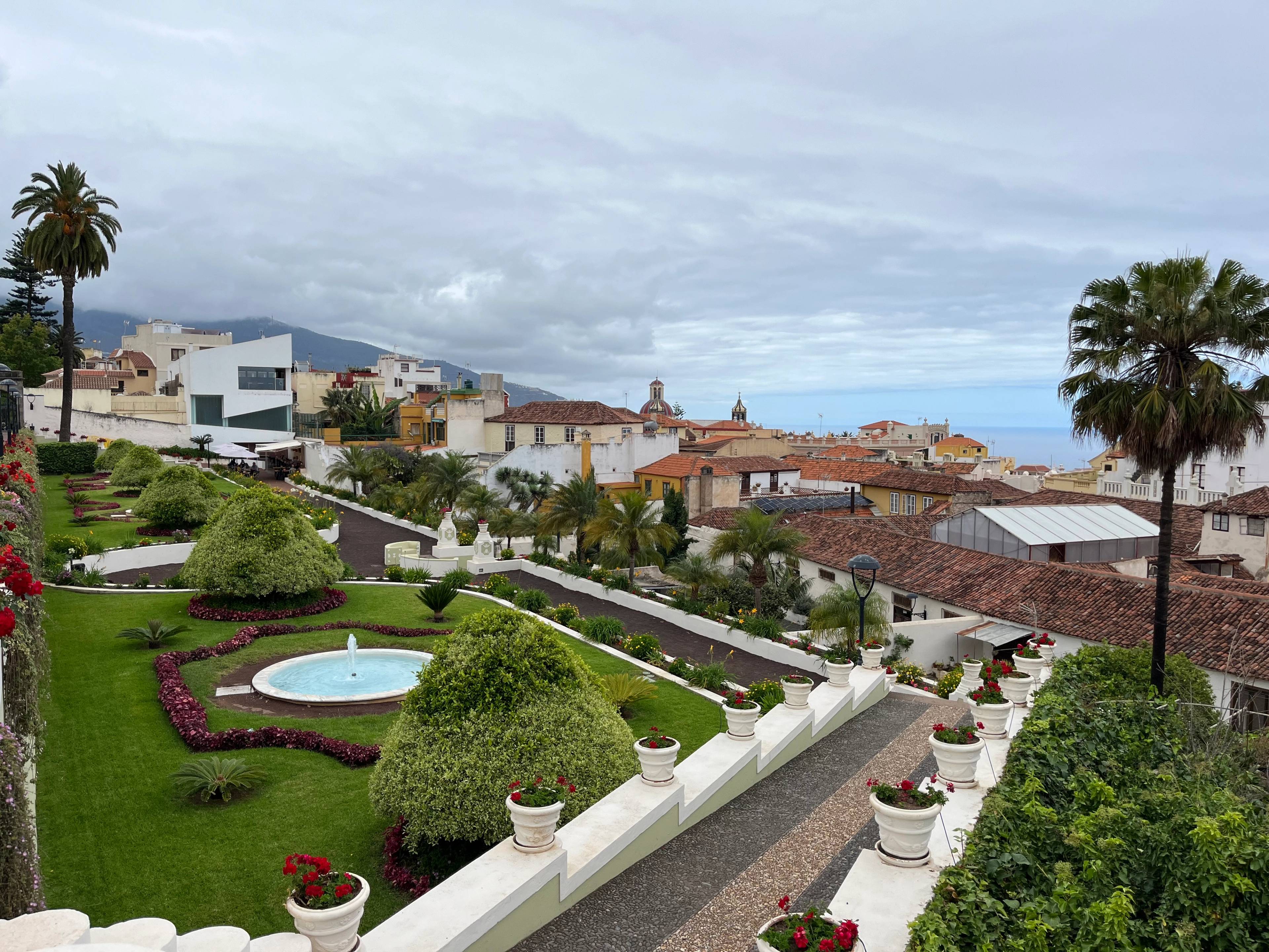Tarasowe ogrody z dekoracyjną roślinnością i fontanną z panoramą miasta w tle.