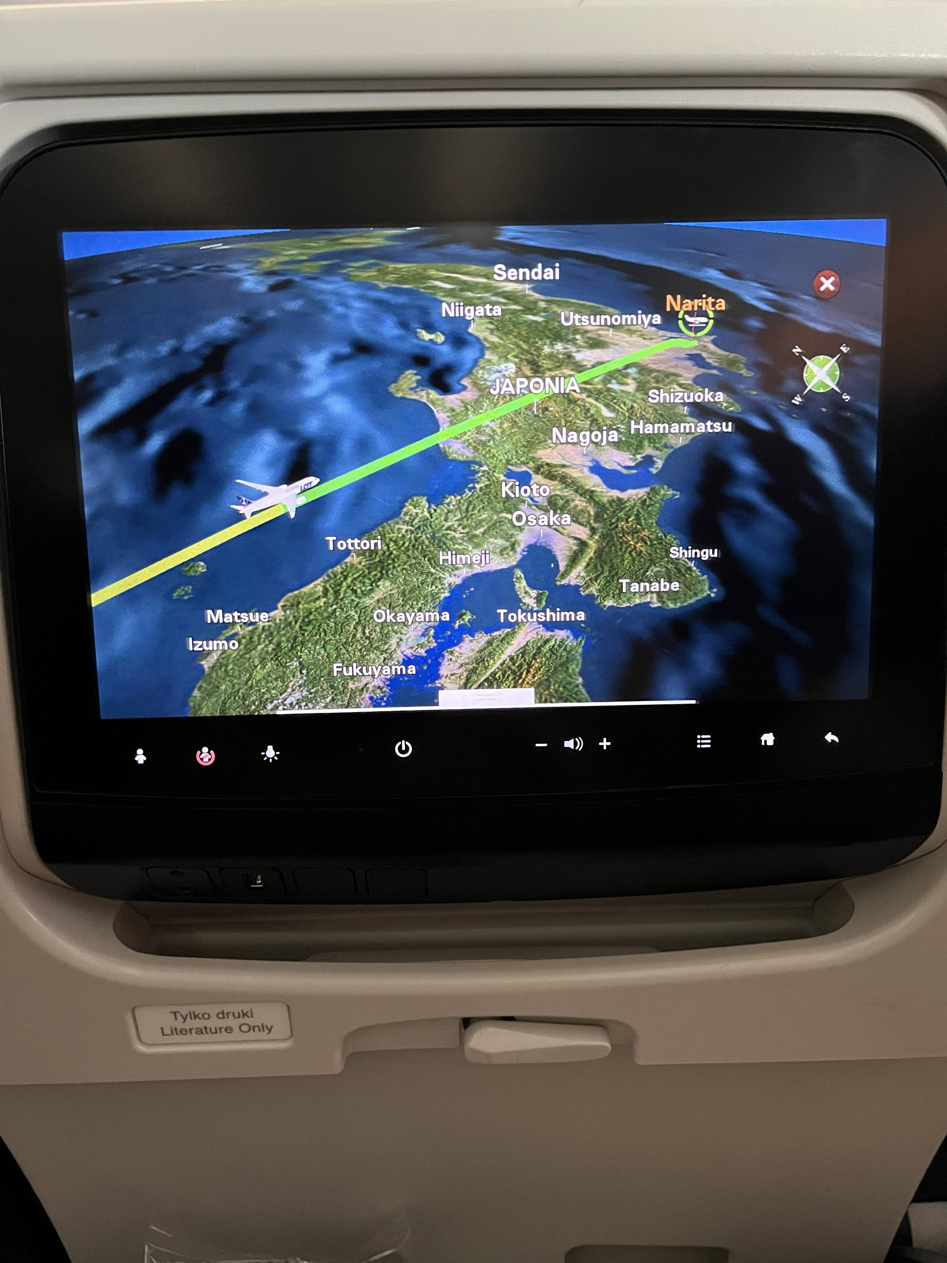 Ekran pokładowego systemu rozrywki przedstawiający mapę Japonii z zaznaczoną trasą lotu i miejscowością Narita jako punktem docelowym podróży.