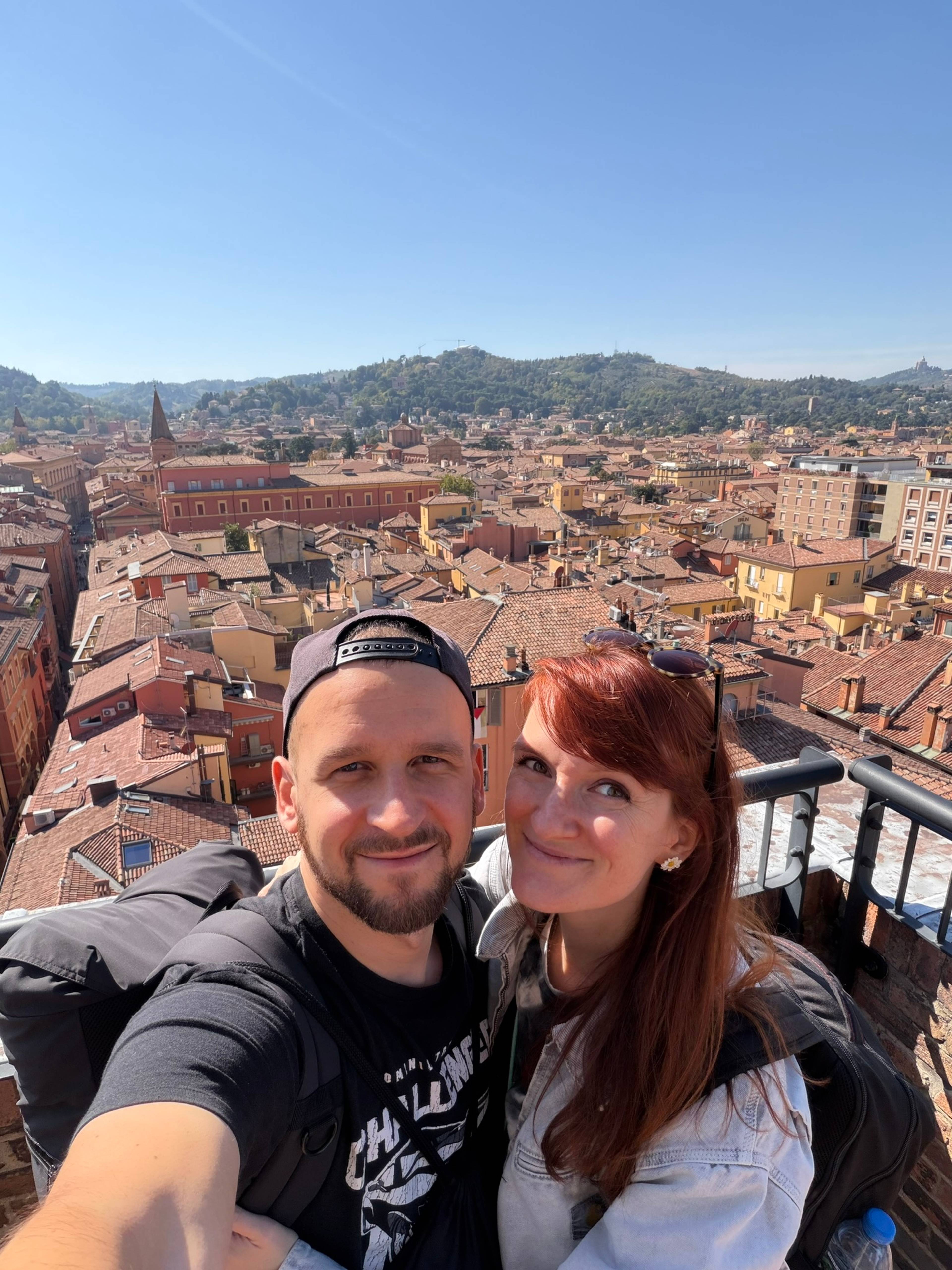 Dwie osoby robią sobie selfie na punkcie widokowym z czerwonymi dachami Bolonii i zielonymi wzgórzami w tle.