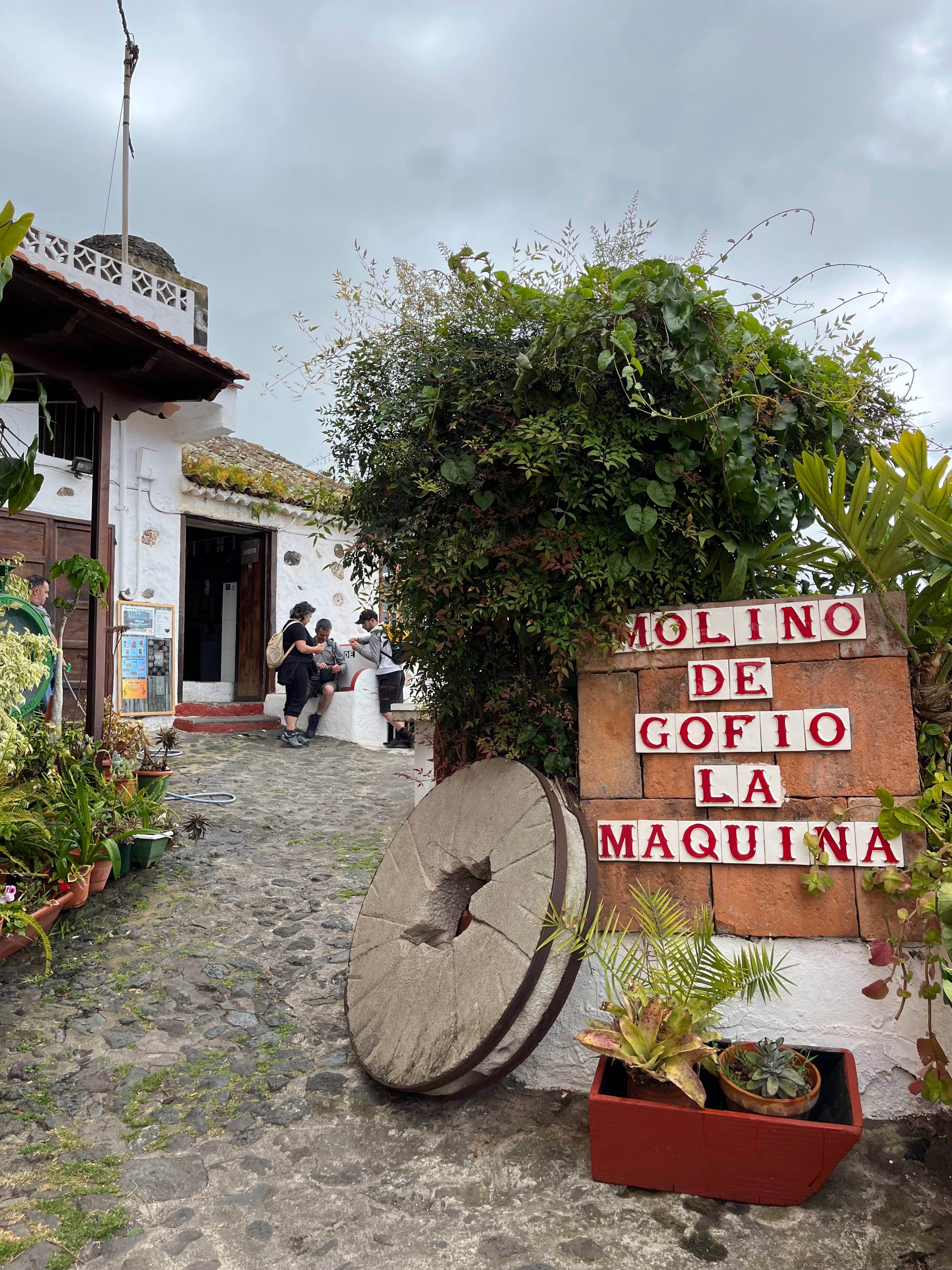 Kamienny młyn i wejście z napisem „Molino de Gofio La Maquina” otoczony roślinami i starymi elementami młyńskimi.
