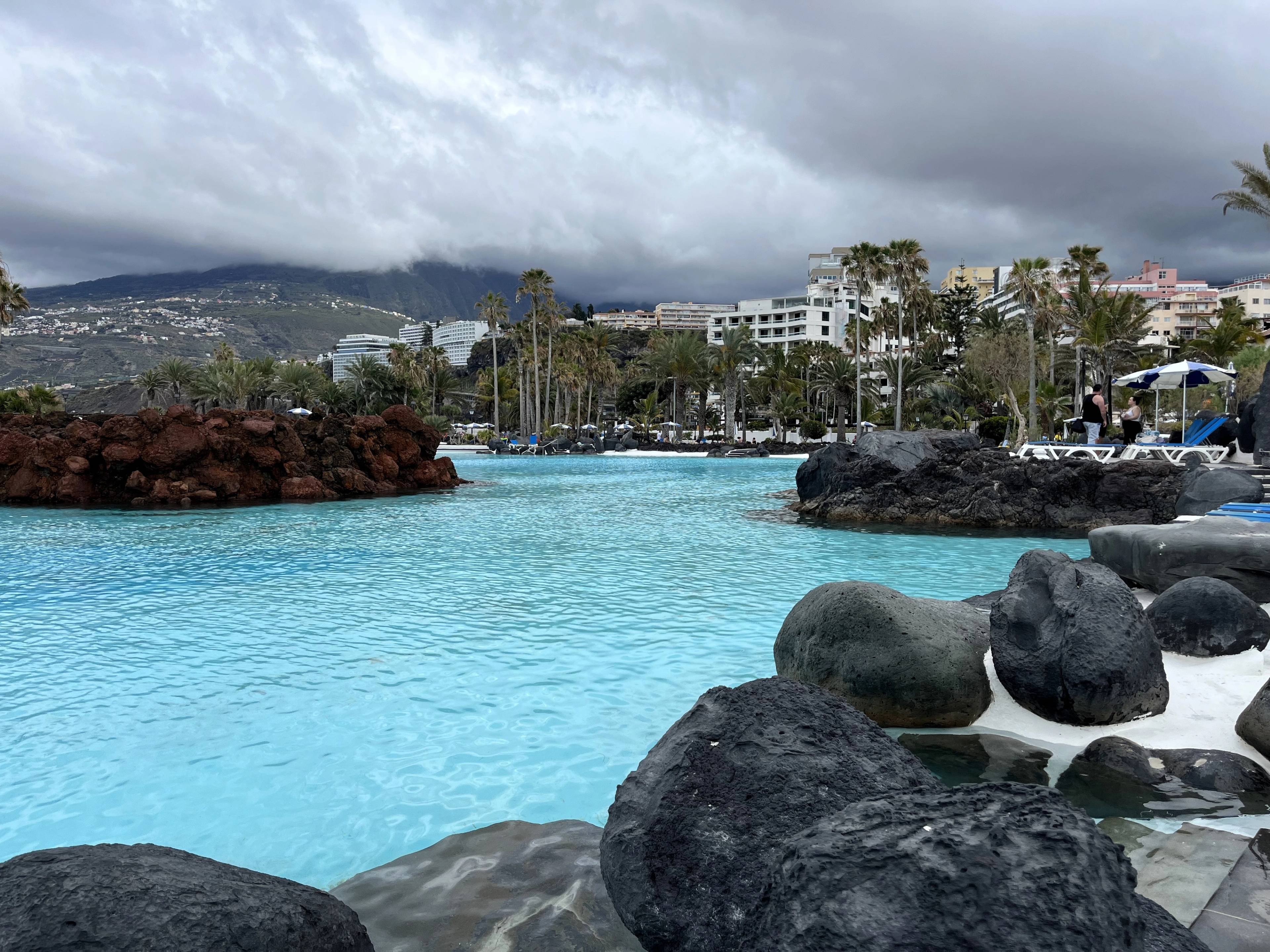 Basen z turkusową wodą otoczony skałami wulkanicznymi, w tle widać zabudowania oraz palmy.