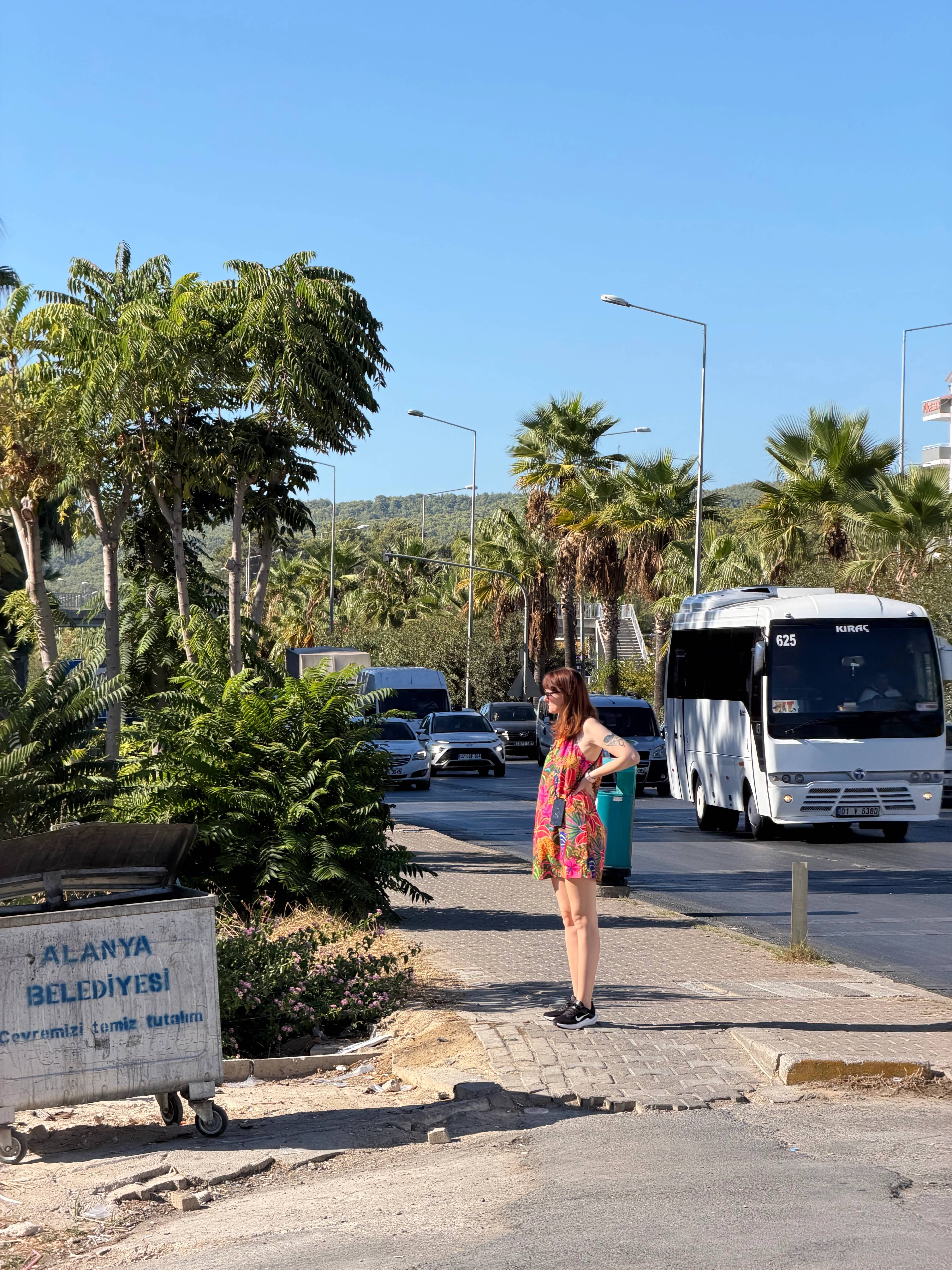 Kobieta stoi na chodniku przy ulicy z palmami i zielonymi krzewami, w tle widoczne samochody i biały autobus na drodze pod jasnym, słonecznym niebem.