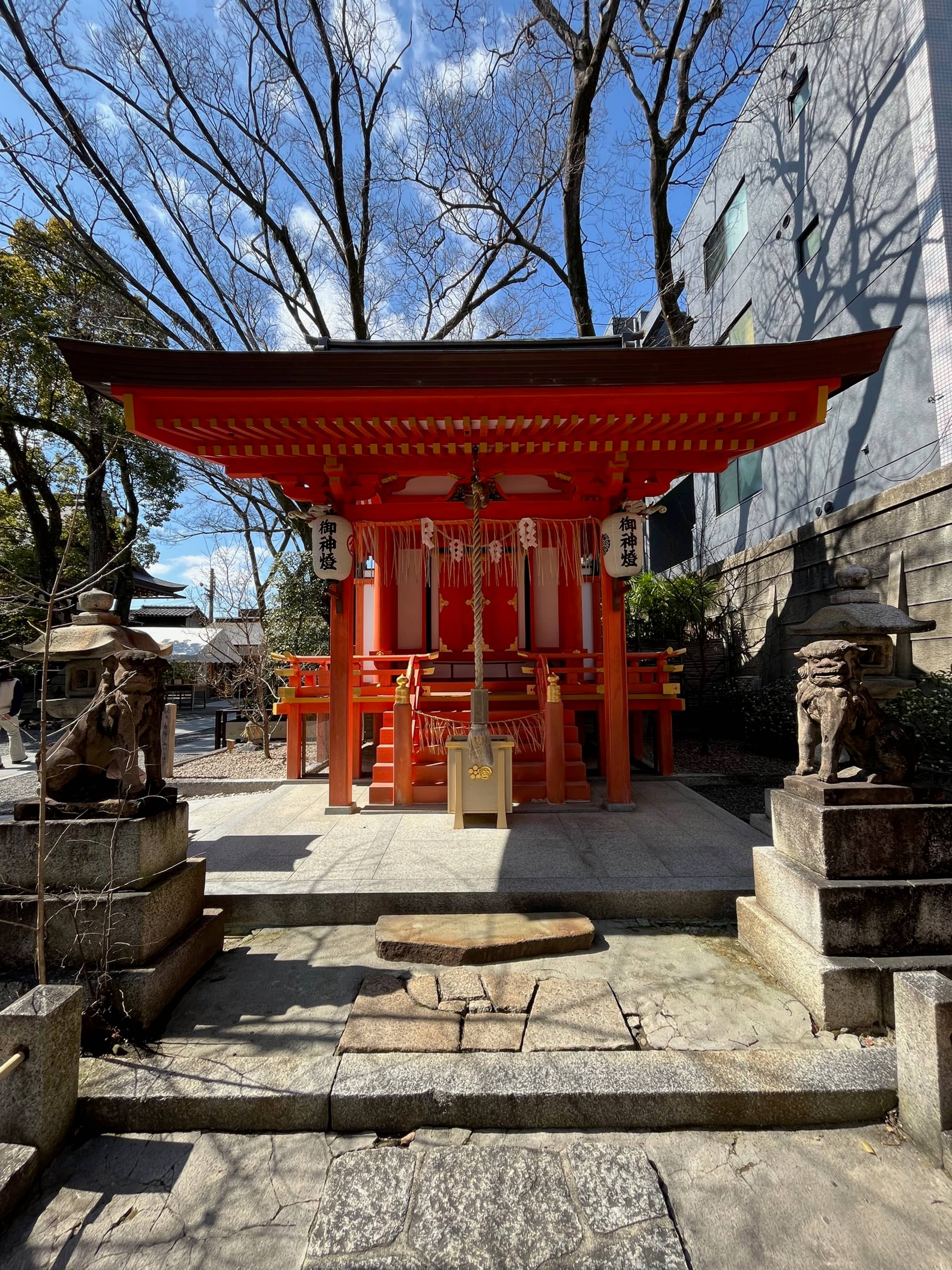 Mała czerwona świątynia shinto stoi na kamiennym podwyższeniu, a po bokach wejścia widoczne są kamienne rzeźby strażników.