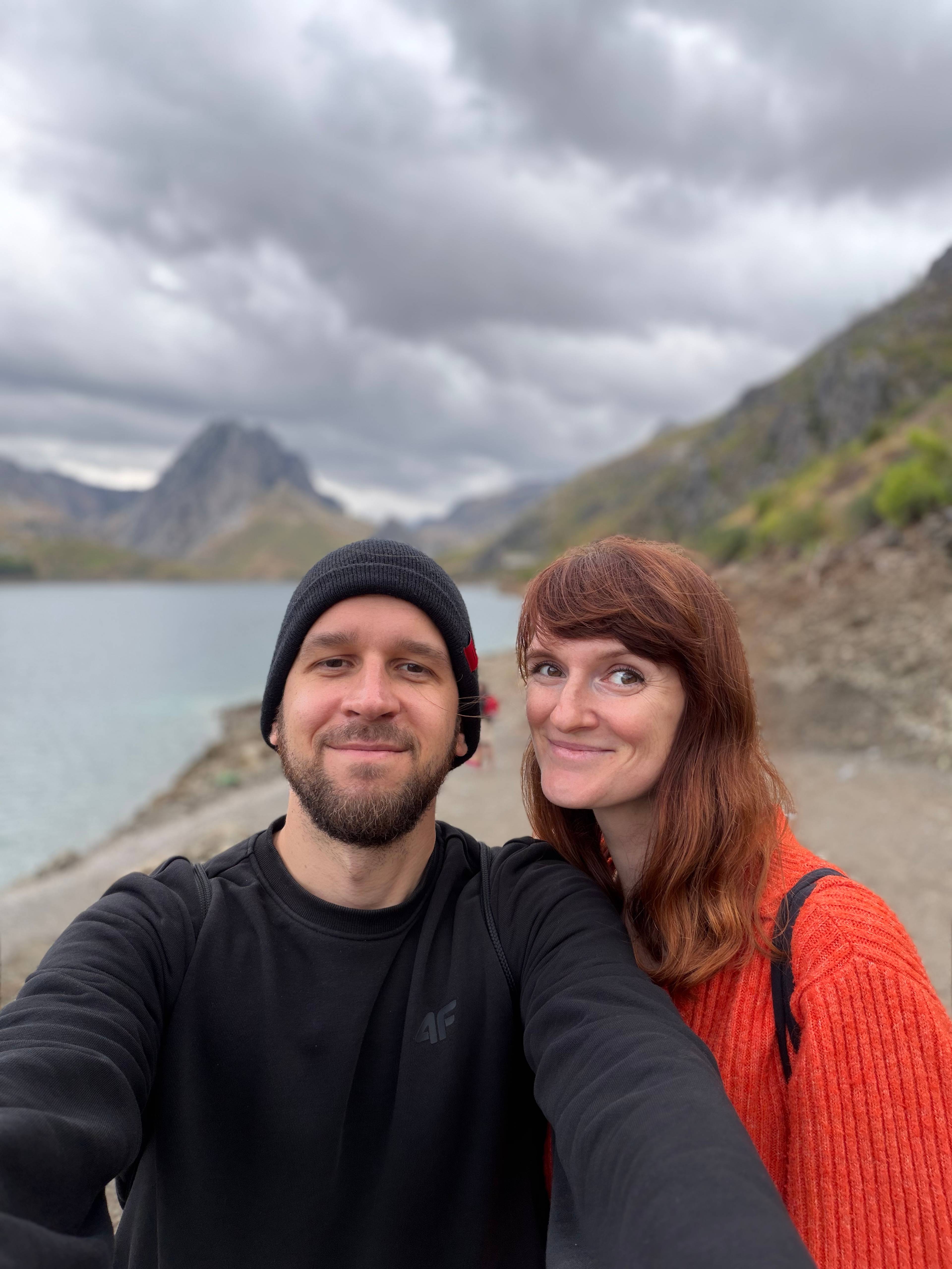 Mężczyzna i kobieta stoją nad brzegiem jeziora z górami w tle pod pochmurnym niebem.