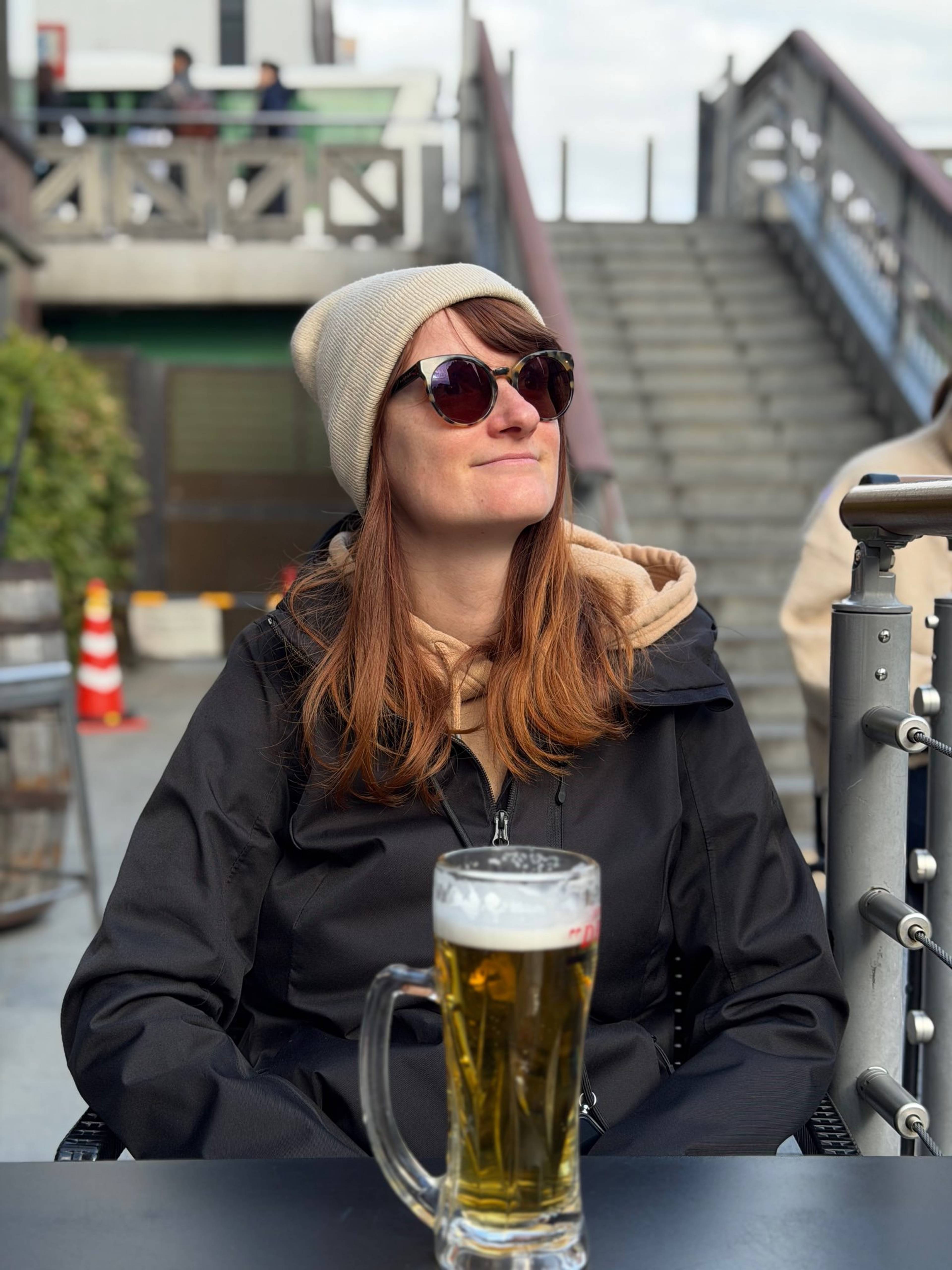 Kobieta w okularach przeciwsłonecznych siedzi przy stoliku na zewnątrz i trzyma kufel jasnego piwa, a w tle widać schody, barierki oraz elementy miejskiej zabudowy.