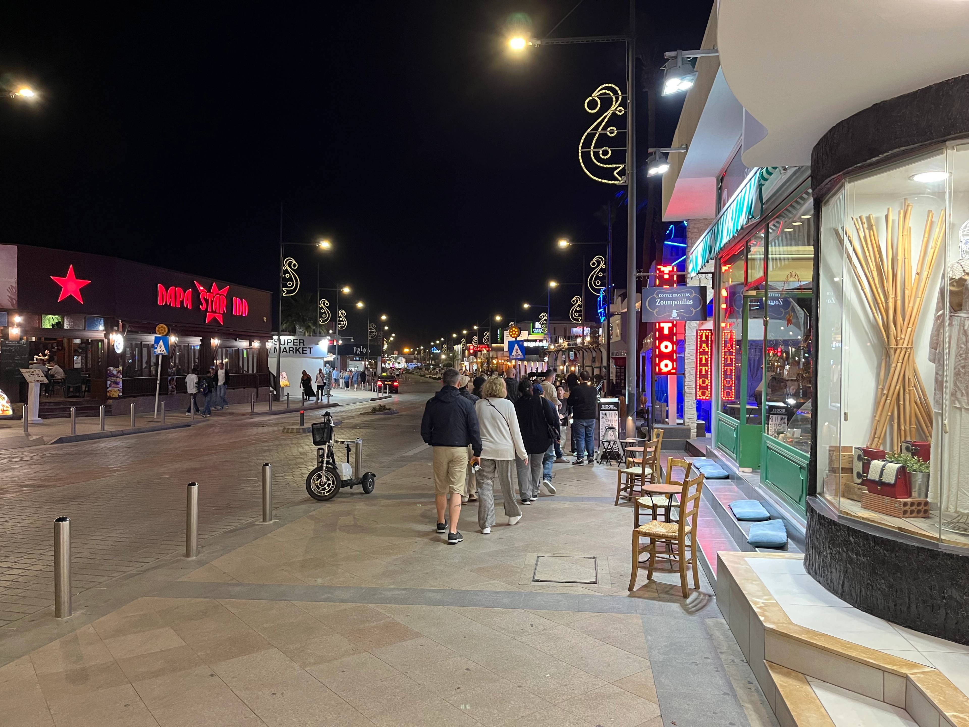 Nocny widok na główną ulicę w Ayia Napa na Cyprze, z kolorowymi neonami, restauracjami i spacerującymi ludźmi.