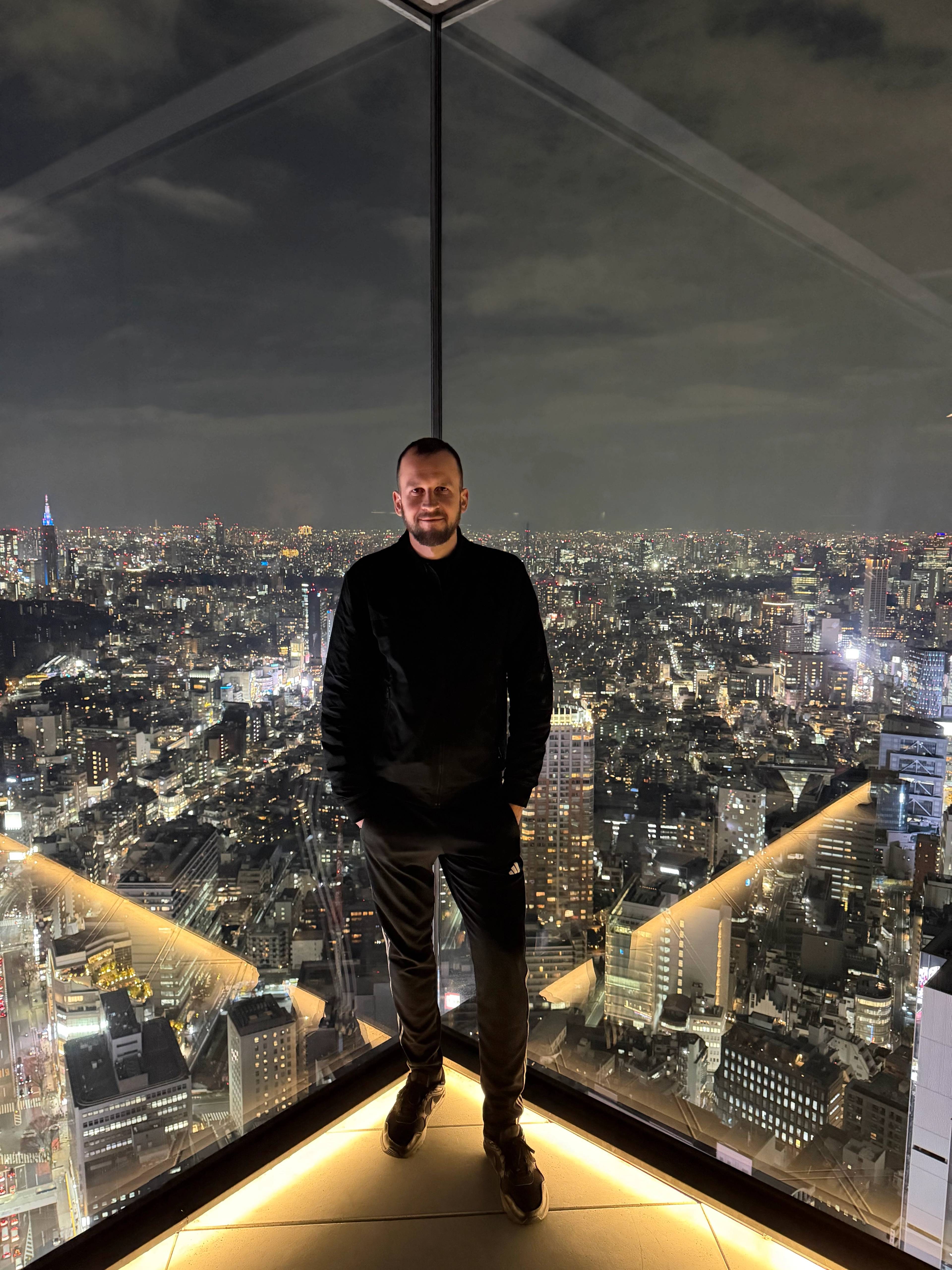 Mężczyzna stojący na tarasie widokowym Shibuya Sky nocą, z panoramicznym widokiem na oświetloną panoramę miasta i drapaczami chmur w tle za jego plecami.
