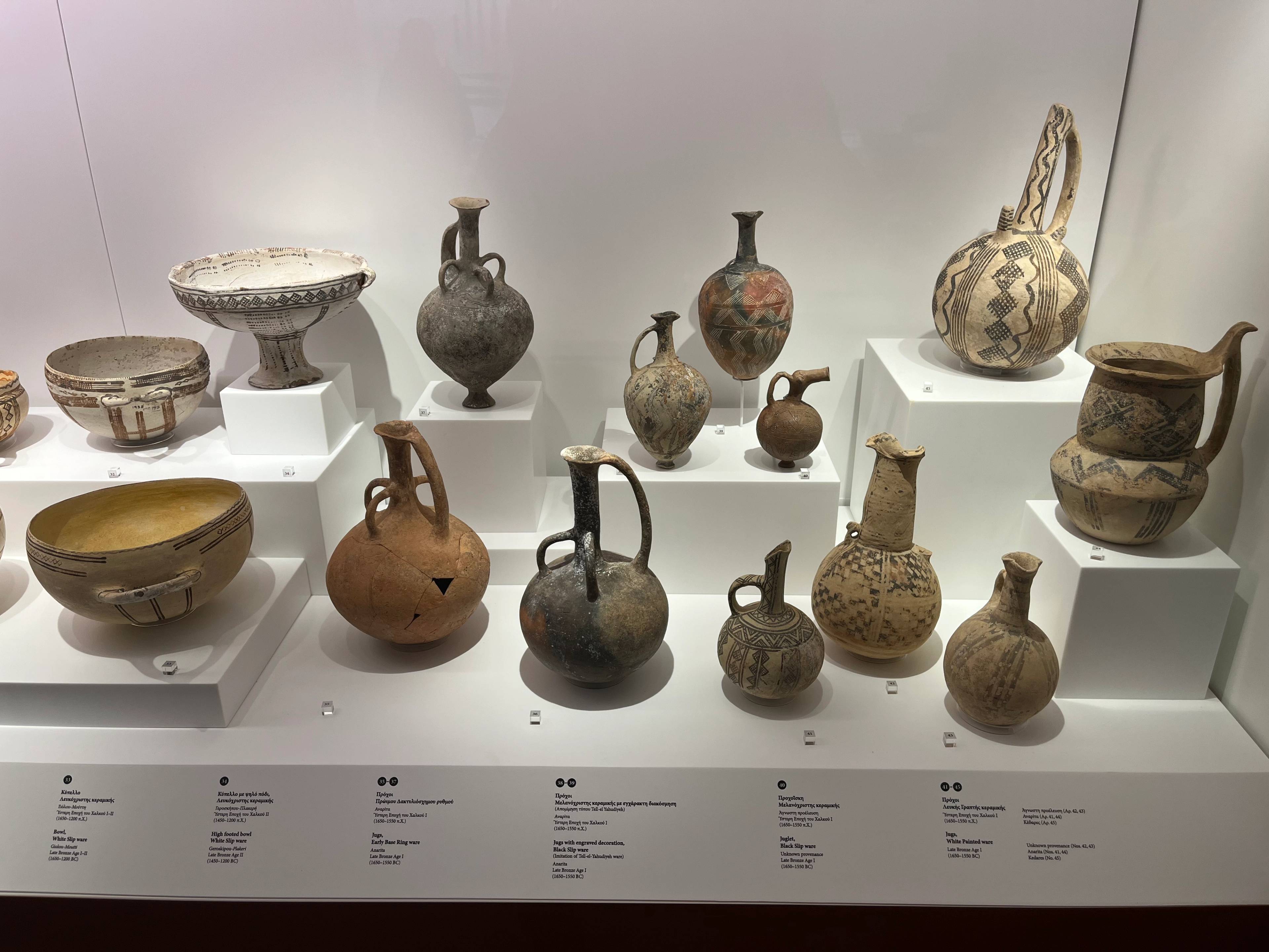 Ekspozycja starożytnych naczyń ceramicznych w muzeum; misy, dzbany i wazony o różnych kształtach i rozmiarach, ustawione na białych podestach z opisami, zdobione wzorami i ornamentami.