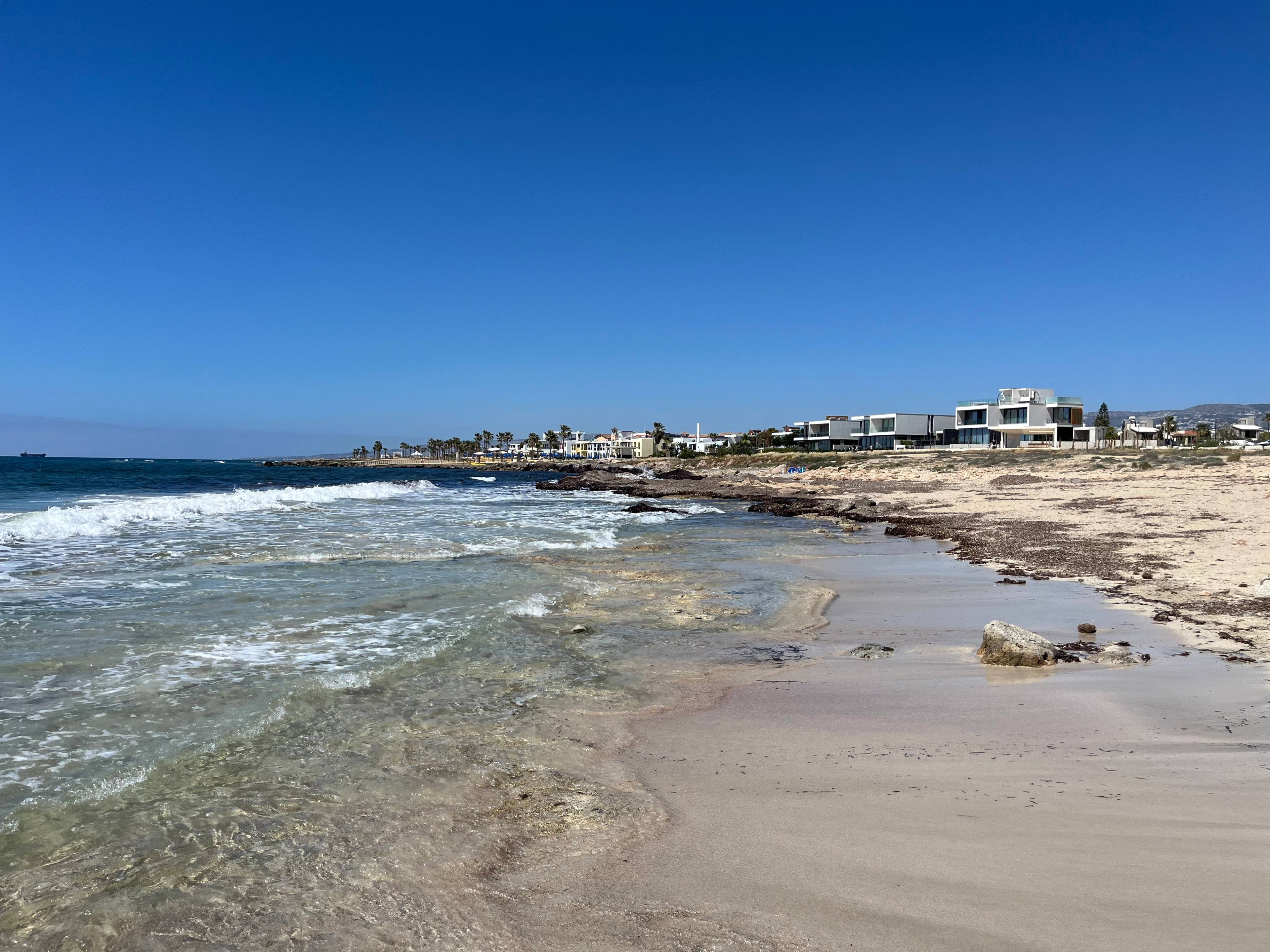 Plaża z jasnym piaskiem, przejrzystą wodą i łagodnymi falami; w tle nowoczesne domy przy samej linii brzegowej, a na brzegu widoczne skały i wodorosty.