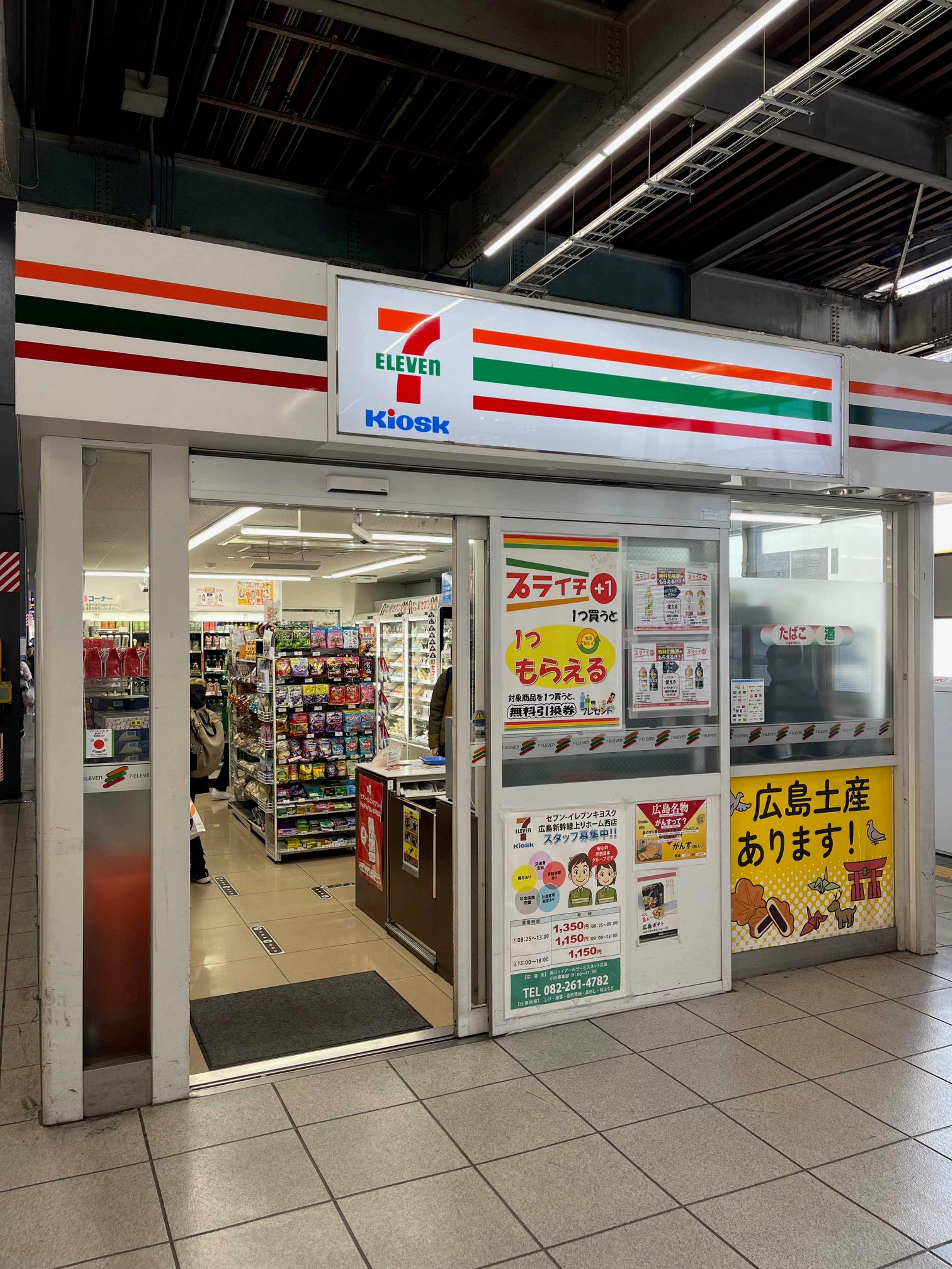 Wejście do sklepu 7‑Eleven na dworcu kolejowym z widocznymi półkami z produktami i japońskimi plakatami informacyjnymi.