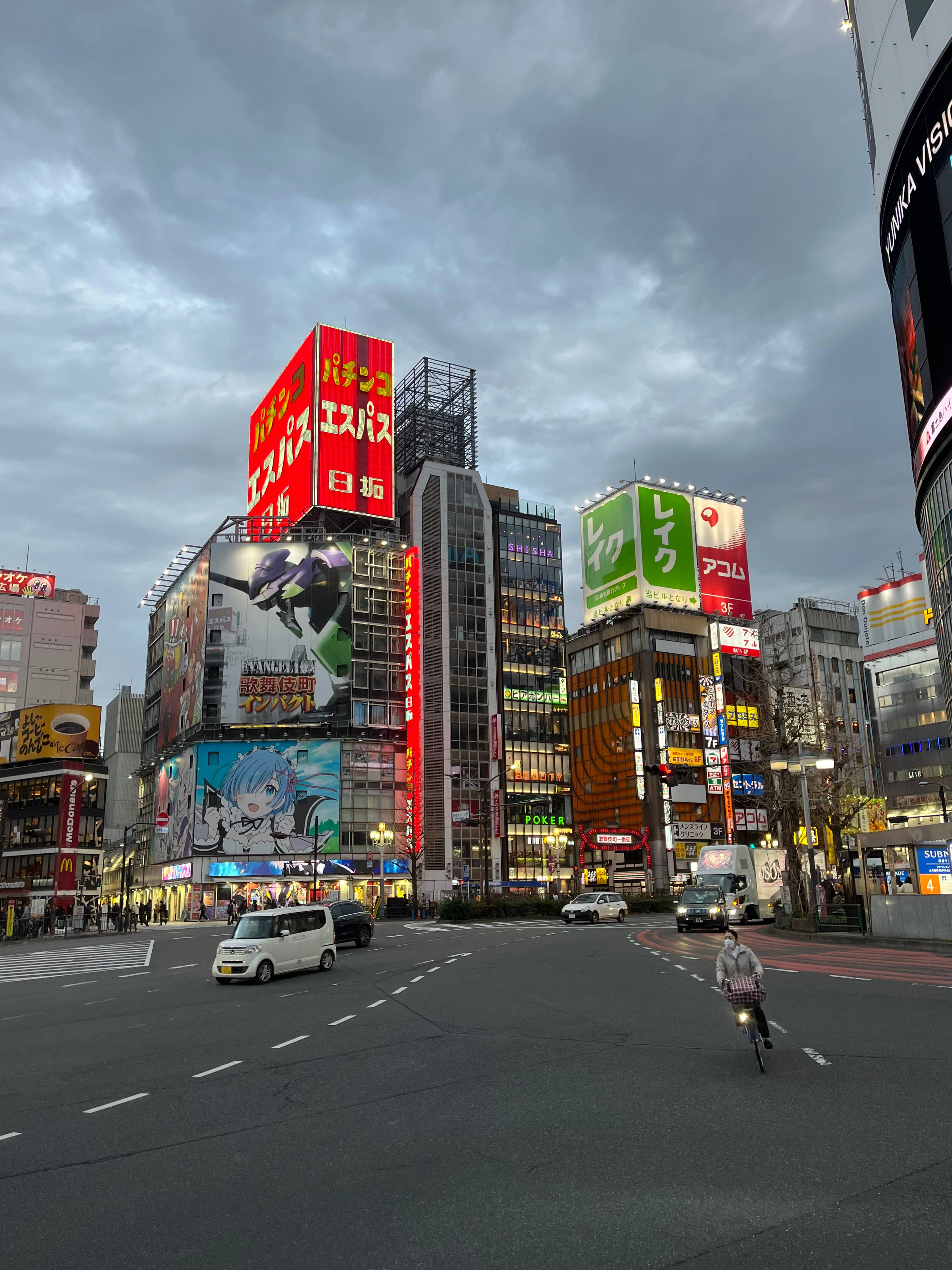 Skrzyżowanie w dzielnicy Shinjuku w Tokio o zmierzchu, z wysokimi budynkami, neonowymi szyldami reklamowymi i ruchem miejskim na ulicy.