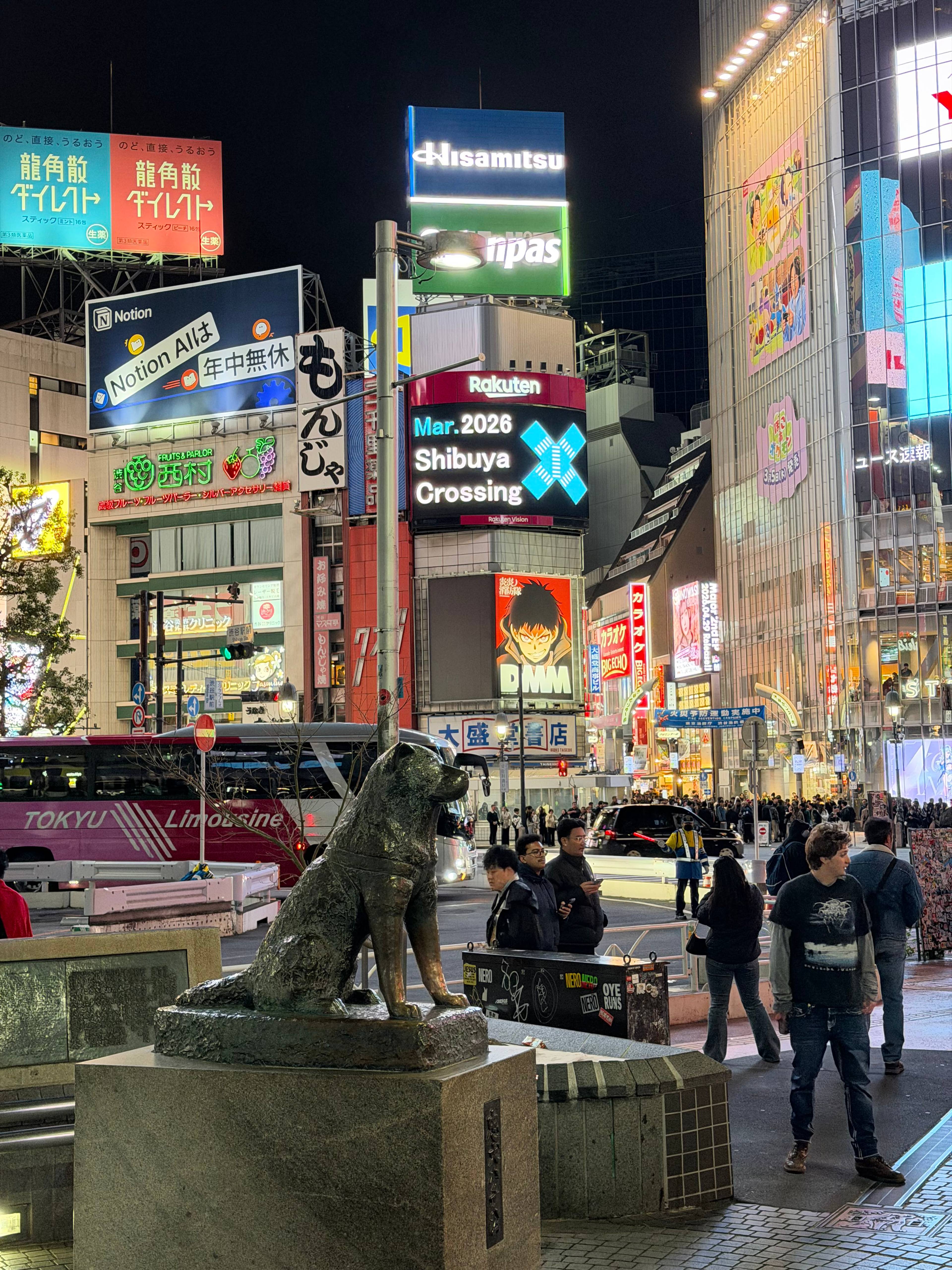Pomnik psa Hachikō przy Shibuya Crossing w Tokio, w tle tłum ludzi i neonowe reklamy nocą.