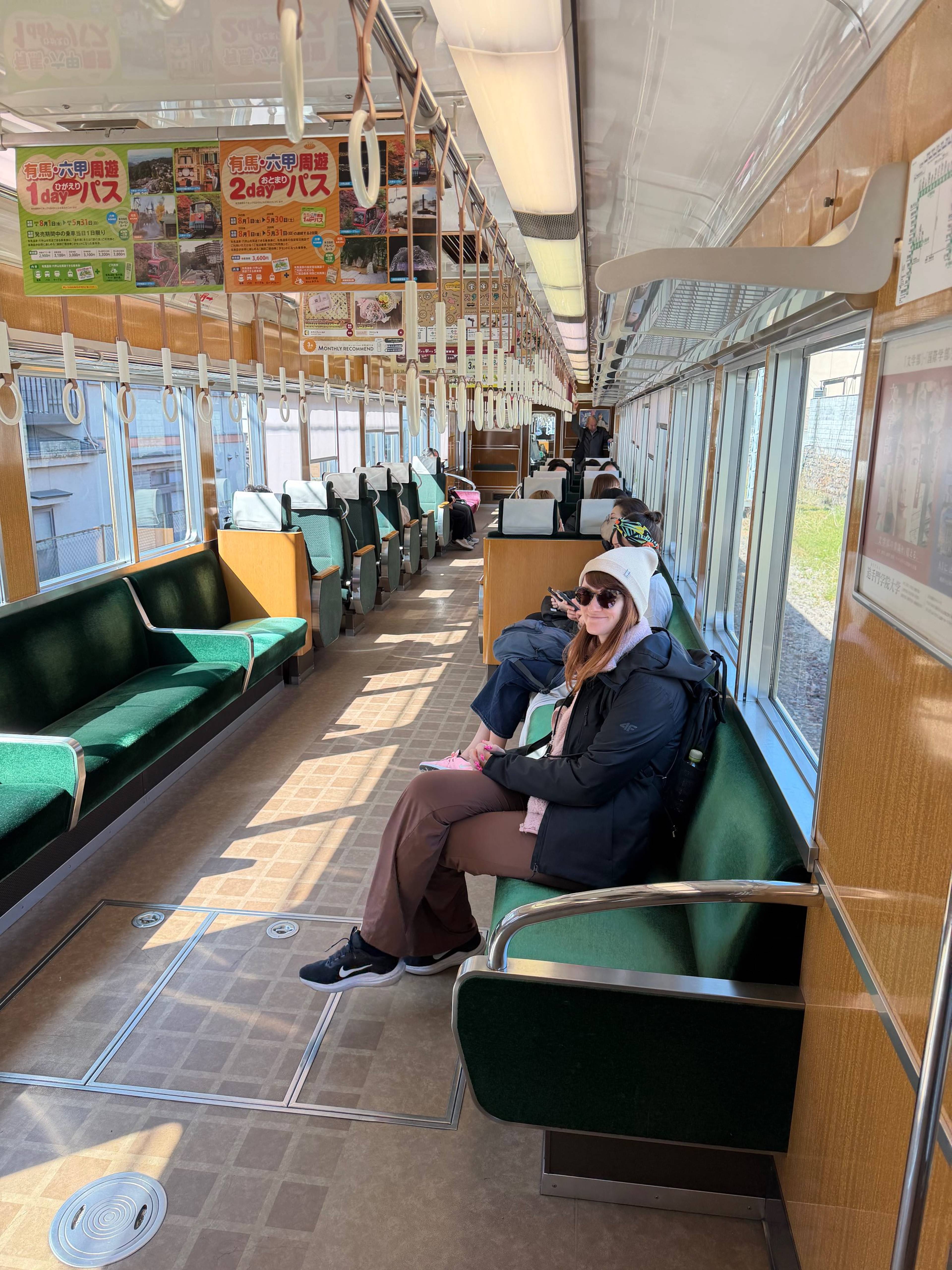 Kobieta siedzi na zielonym siedzeniu wewnątrz japońskiego pociągu lokalnego, a wzdłuż wagonu widać rzędy miejsc, uchwyty do trzymania oraz plakaty informacyjne nad oknami.