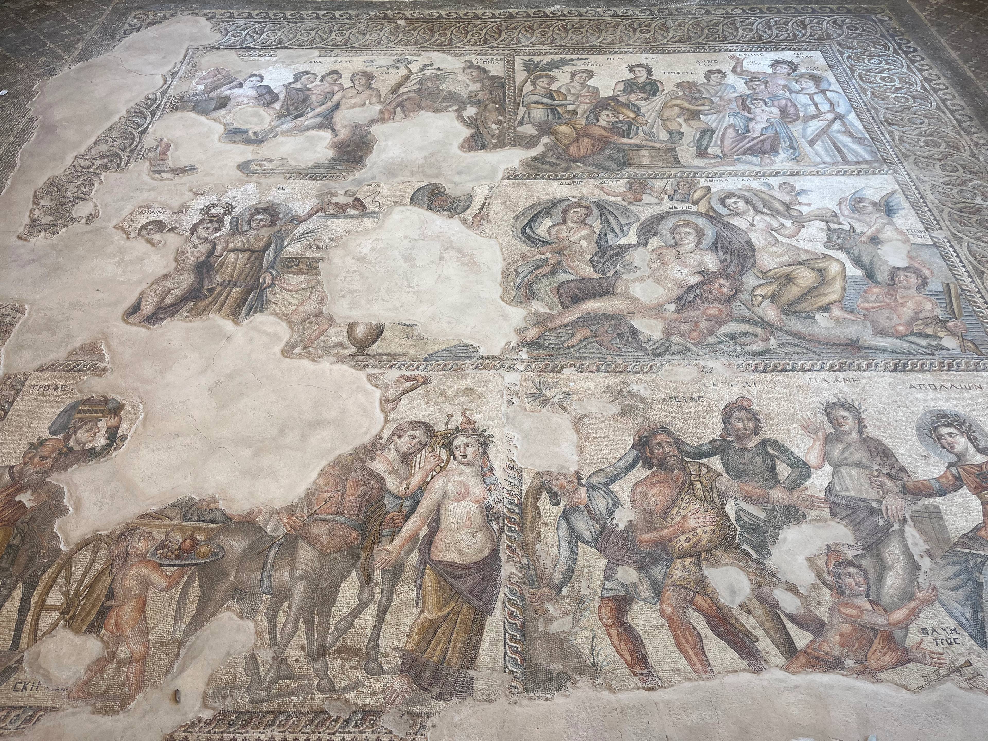 Starożytny mozaikowy panel przedstawiający sceny z udziałem ludzkich postaci, prawdopodobnie o tematyce mitologicznej lub historycznej; mozaika częściowo uszkodzona, z fragmentami wyblakłymi lub brakującymi.