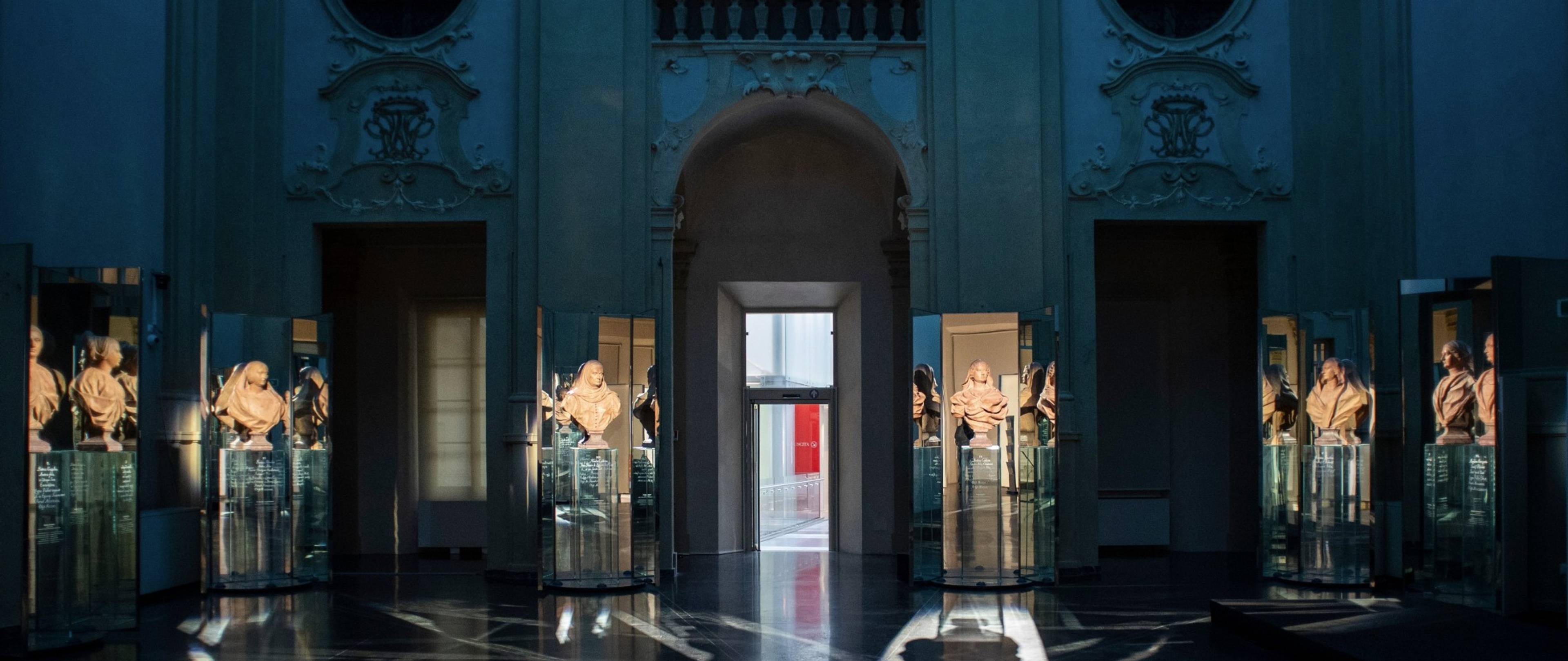 Wnętrze muzeum w Palazzo Pepoli z popiersiami ustawionymi w przeszklonych gablotach i odbiciami światła na ciemnej posadzce.