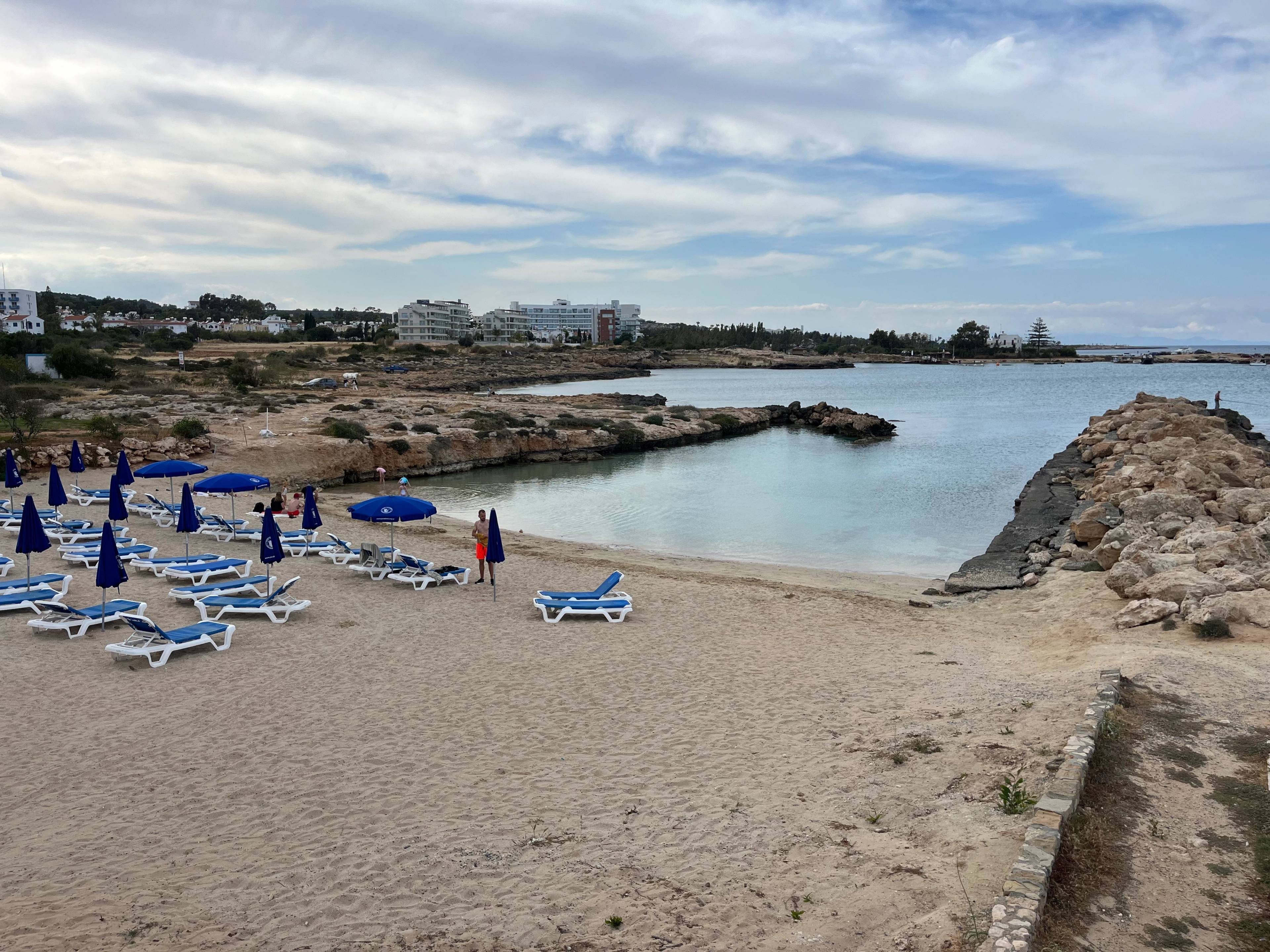 Piaszczysta plaża Glyfades w Protaras na Cyprze z rzędem niebieskich leżaków i parasoli, spokojną zatoką oraz hotelami w tle.