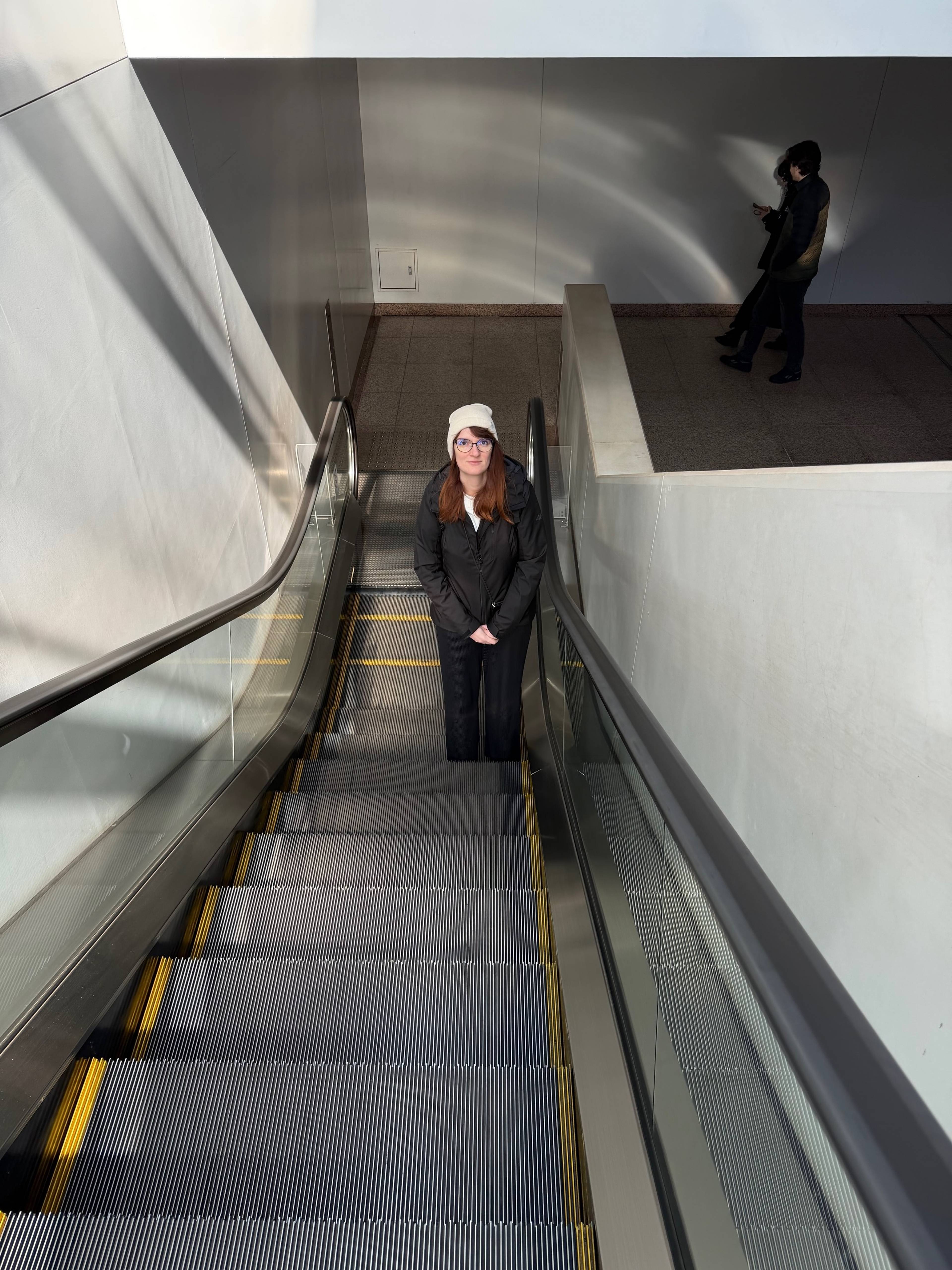 Kobieta stoi na schodach ruchomych w nowoczesnym przejściu podziemnym, a jasne ściany i metalowe balustrady prowadzą wzrok w dół przestrzeni.