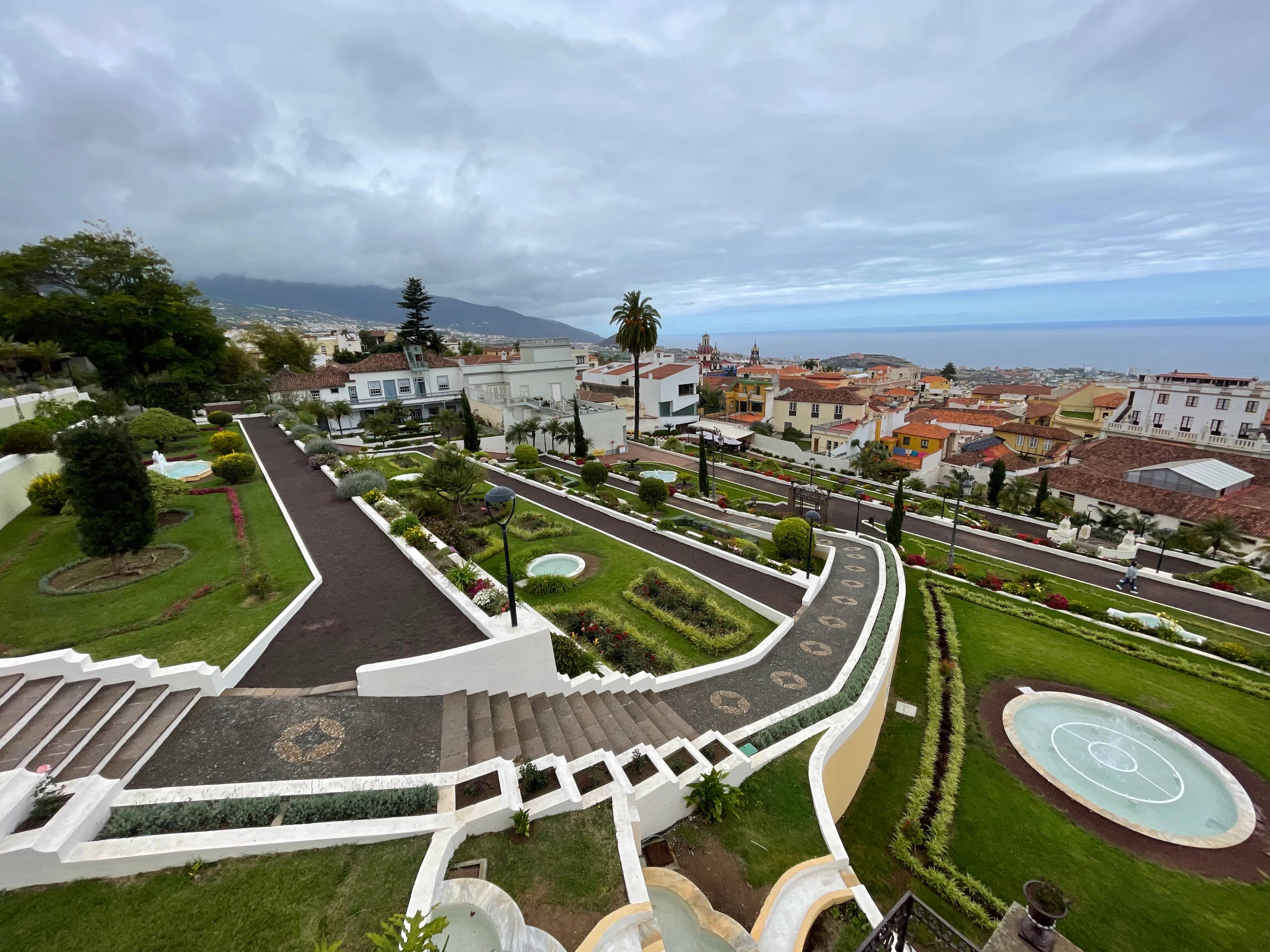 Tarasowe ogrody z fontannami i alejkami z widokiem na miasto i ocean w oddali.