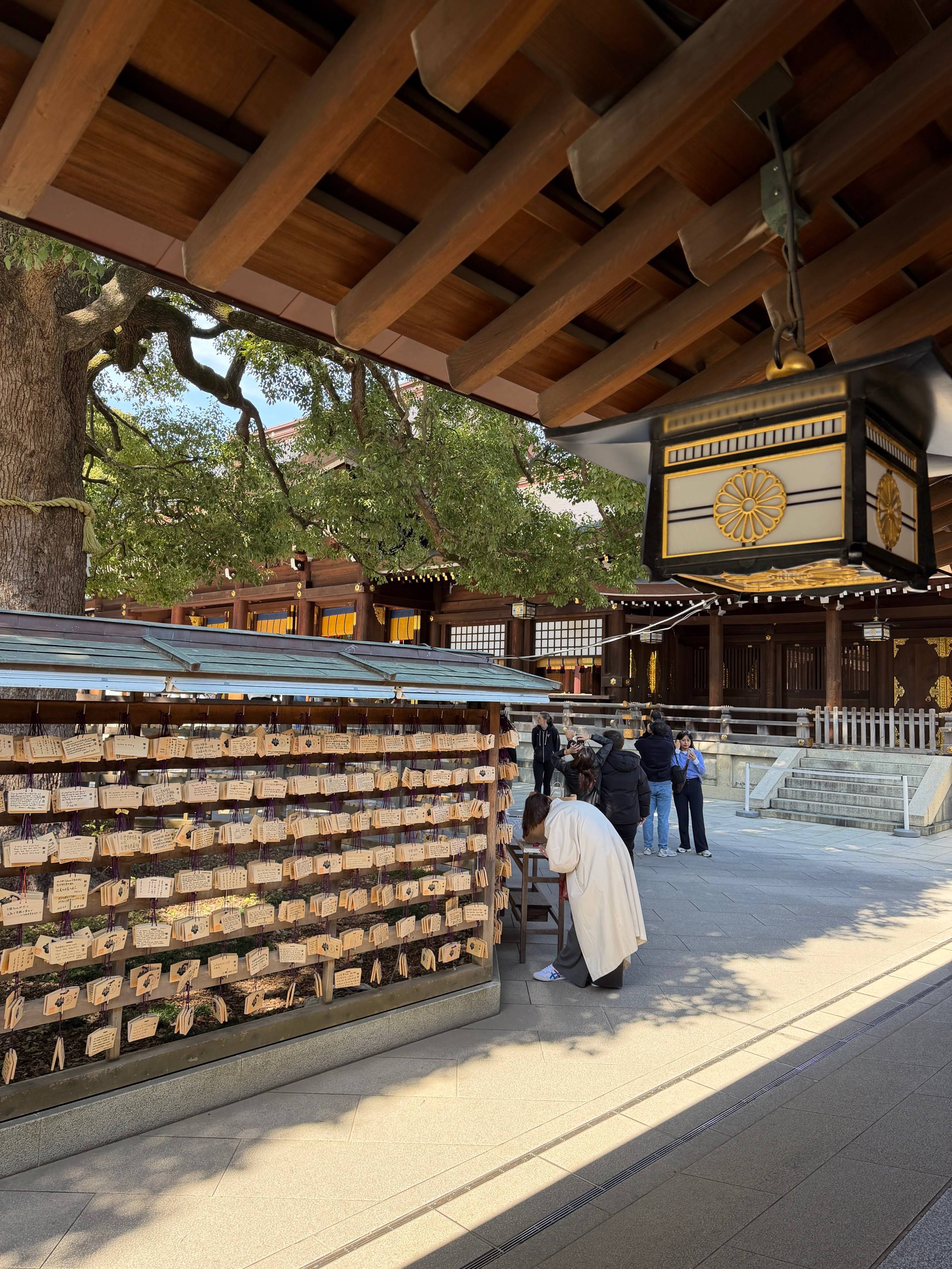 Dziedziniec japońskiej świątyni z drewnianymi budynkami, dużą lanterną i stojakiem z ema, przy którym odwiedzający oglądają zawieszone drewniane tabliczki.