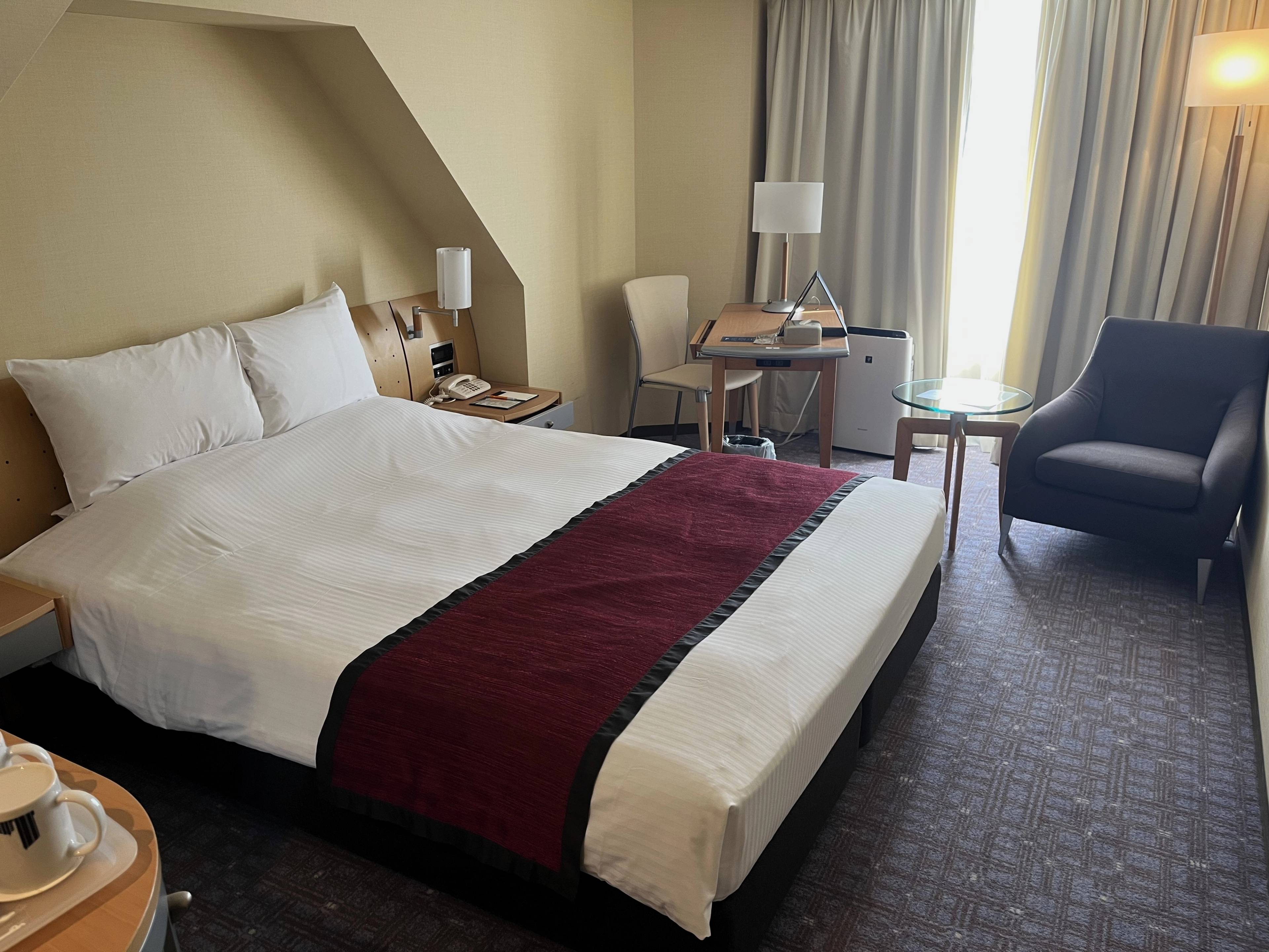 Pokój hotelowy w Tokio z dużym łóżkiem, czerwonym bieżnikiem, biurkiem, fotelem i zasłoniętym oknem.