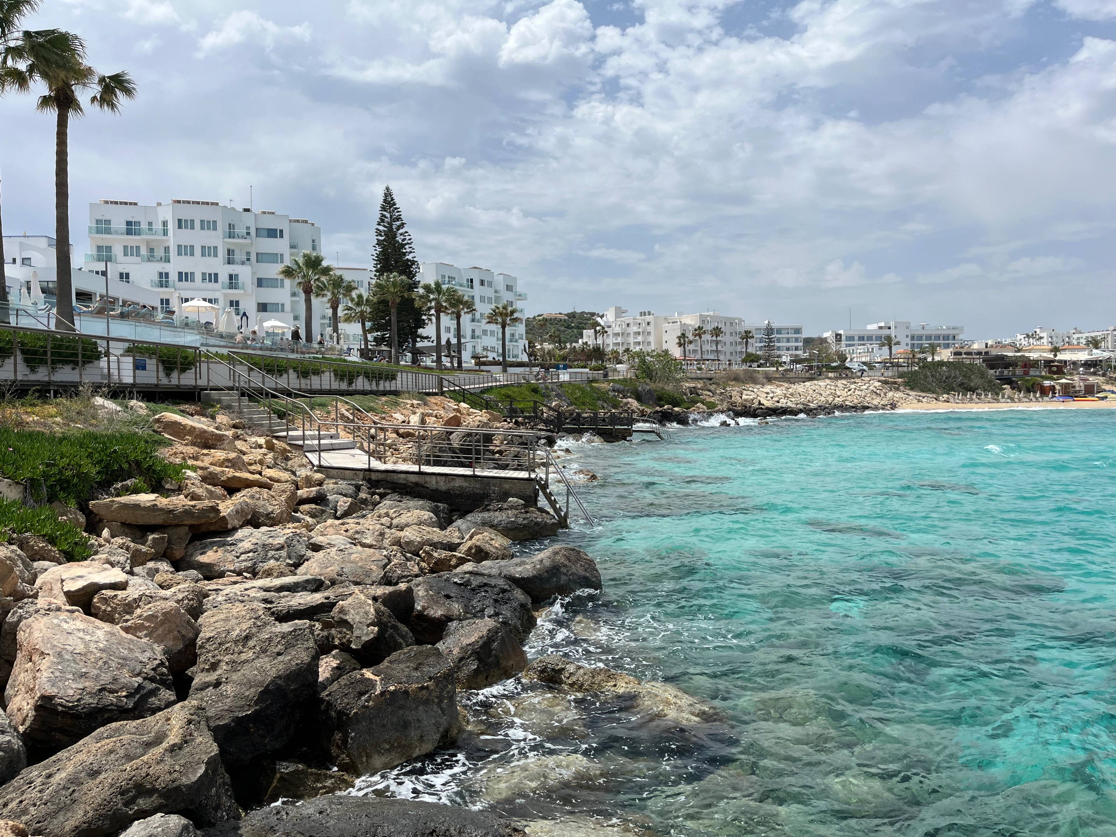 Widok na skaliste wybrzeże Protaras na Cyprze z turkusową wodą i hotelami w tle; poniżej drewniana promenada prowadząca wzdłuż wybrzeża.