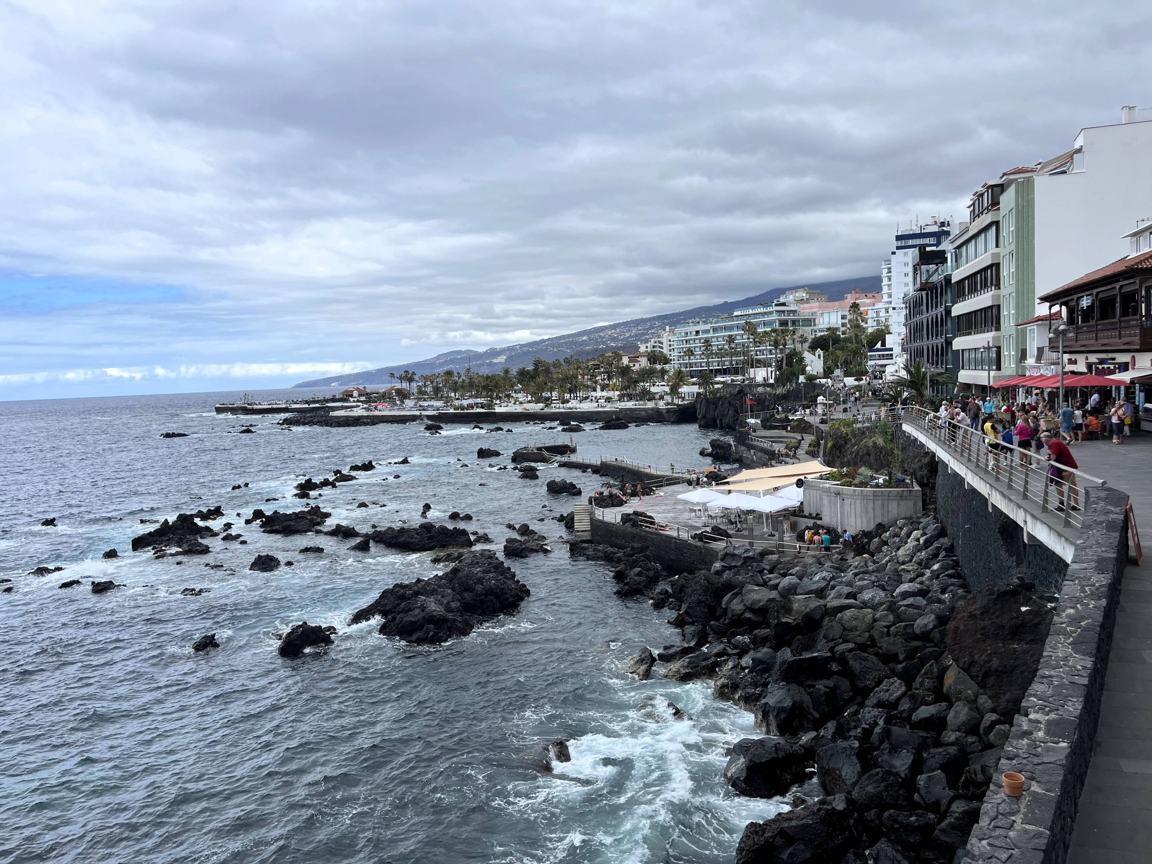 Widok na nadmorską promenadę, budynki hoteli i restauracji, a na większej części kadru morze z kamieniami wulkanicznymi.