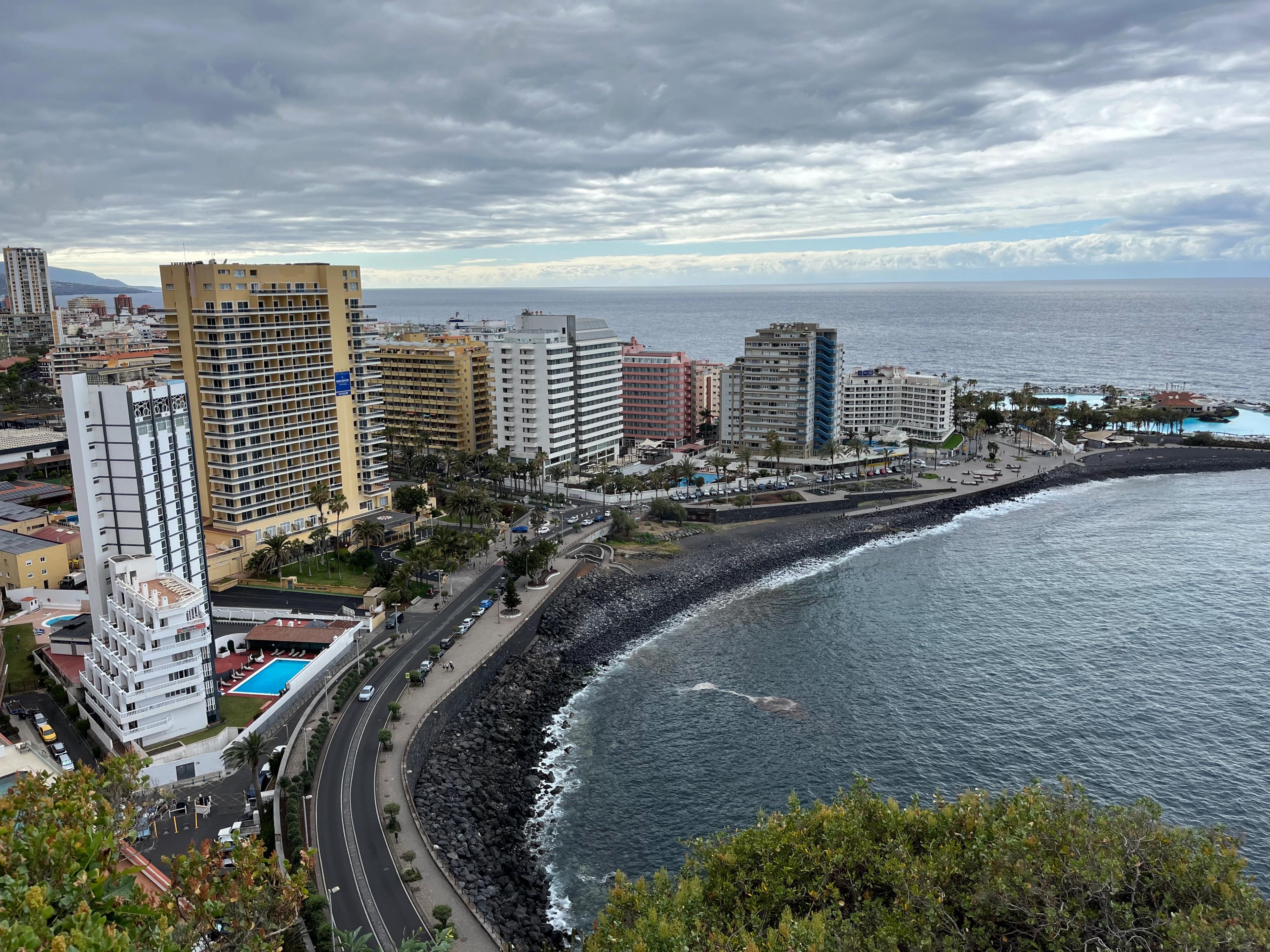 Nadmorska panorama, widok z góry na długą, czarną, kamienistą plażę oraz wysokie kolorowe budynki - głównie wielopiętrowe hotele.