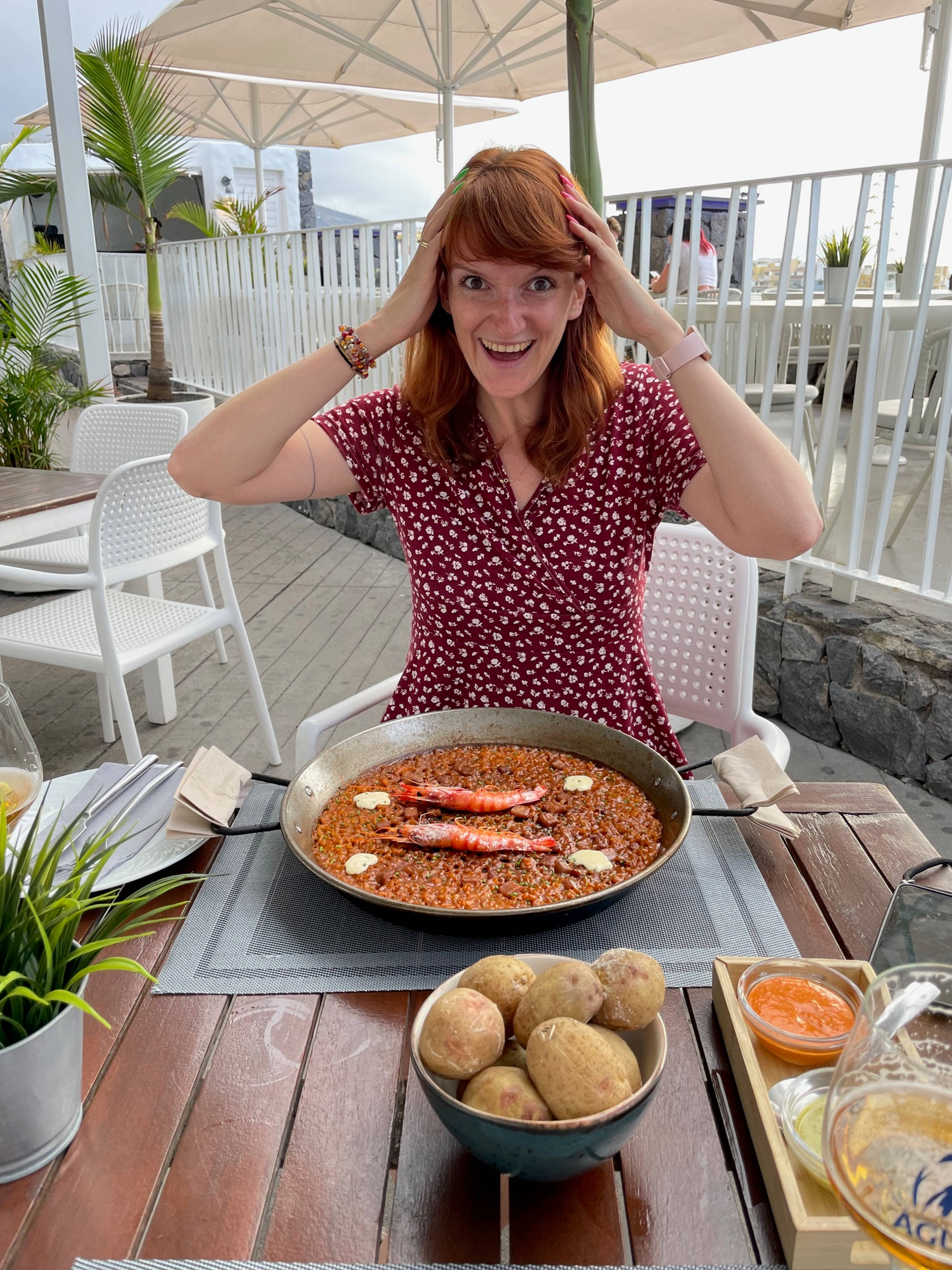 Kobieta siedzi przy stole z dużą paellą i ziemniakami, trzyma się za głowę i jest zaskoczona, że ma tyle do zjedzenia,