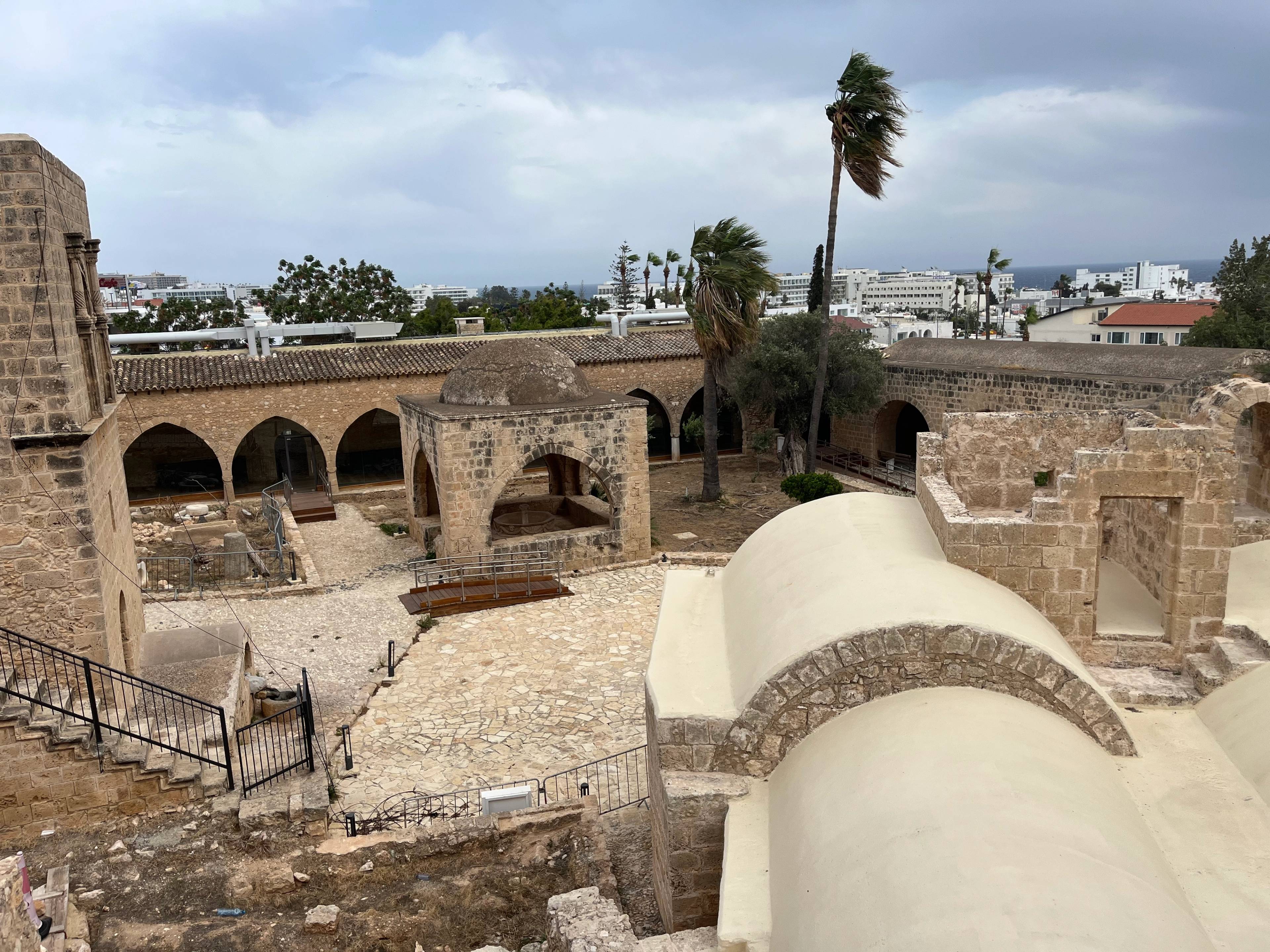 Kamienny dziedziniec klasztoru w Ayia Napa na Cyprze, otoczony arkadami i zabytkowymi murami, z widokiem na miasto w tle.