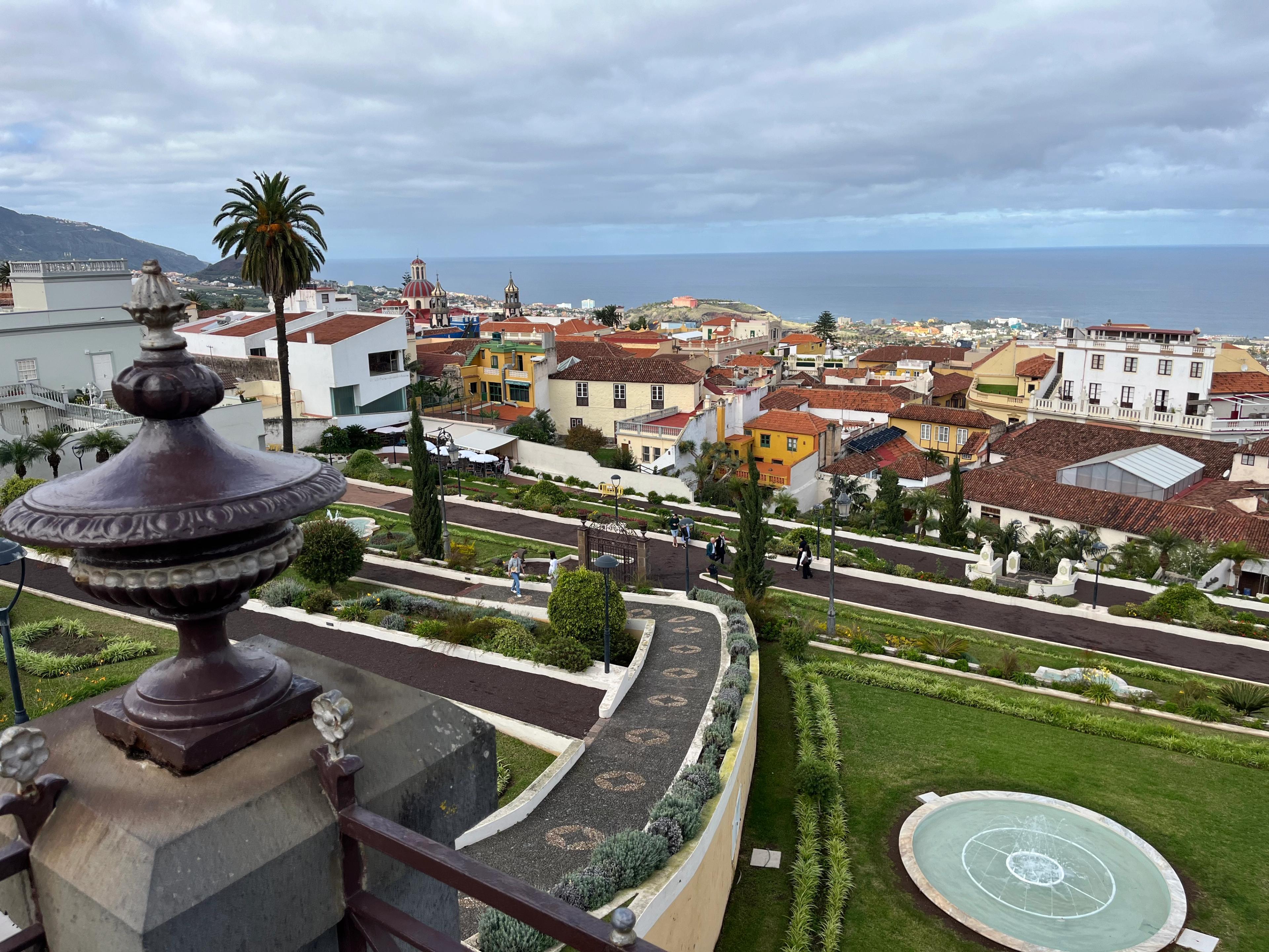 Widok na ogrody oraz miasto z kolorowymi kamienicami i oceanem w tle. Na pierwszym planie wieżyczka zdobiąca brzeg balustrady. 