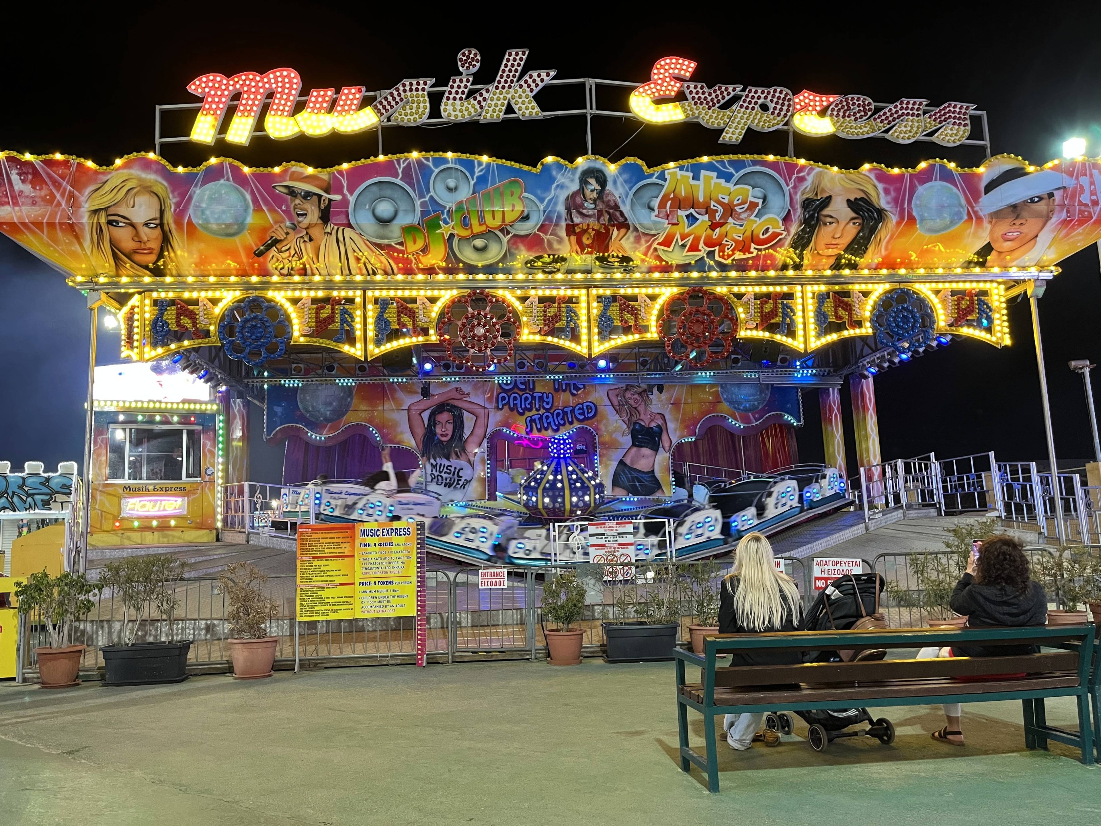 Atrakcja Music Express w wesołym miasteczku Ayia Napa na Cyprze, z kolorową iluminacją i ławkami przed wejściem.