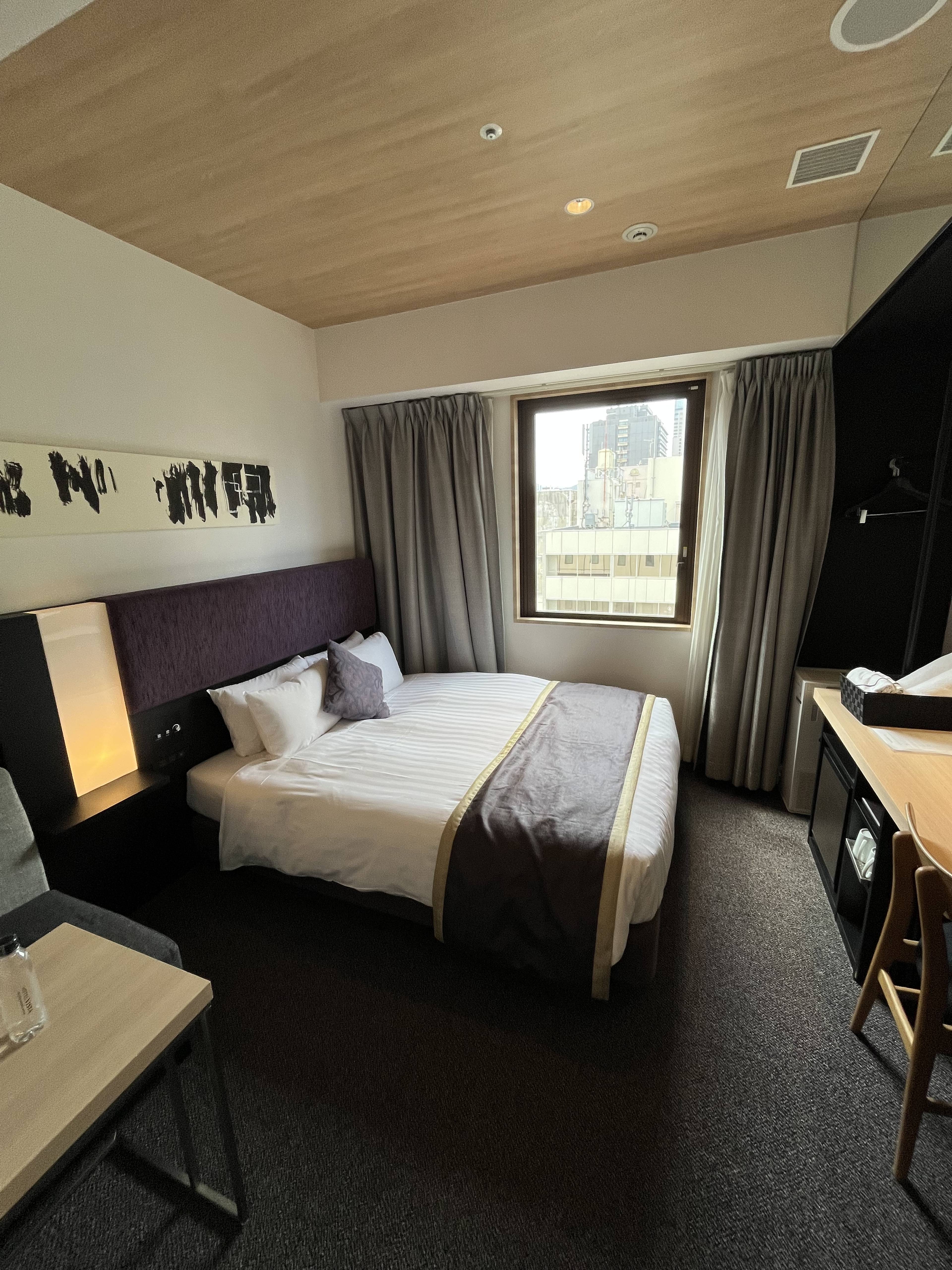 Pokój hotelowy z dwuosobowym łóżkiem, szarą narzutą, biurkiem oraz oknem zasłoniętym ciemnoszarymi zasłonami.