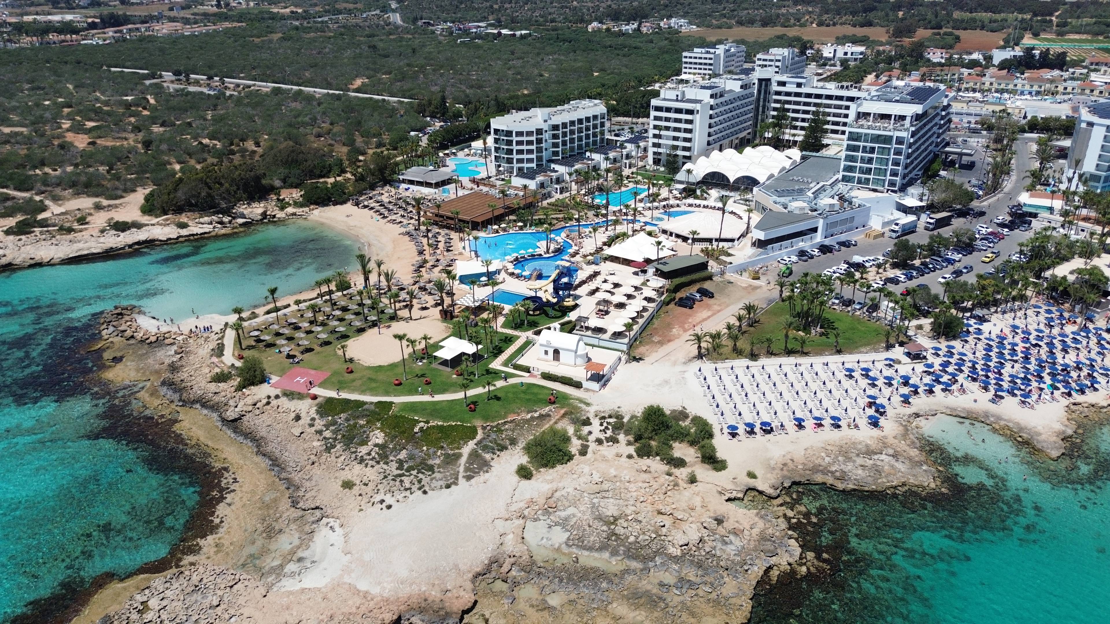 Widok z lotu ptaka na kompleks hotelowy przy plaży w Ayia Napa na Cyprze, z basenami, leżakami i turkusowym morzem w tle.