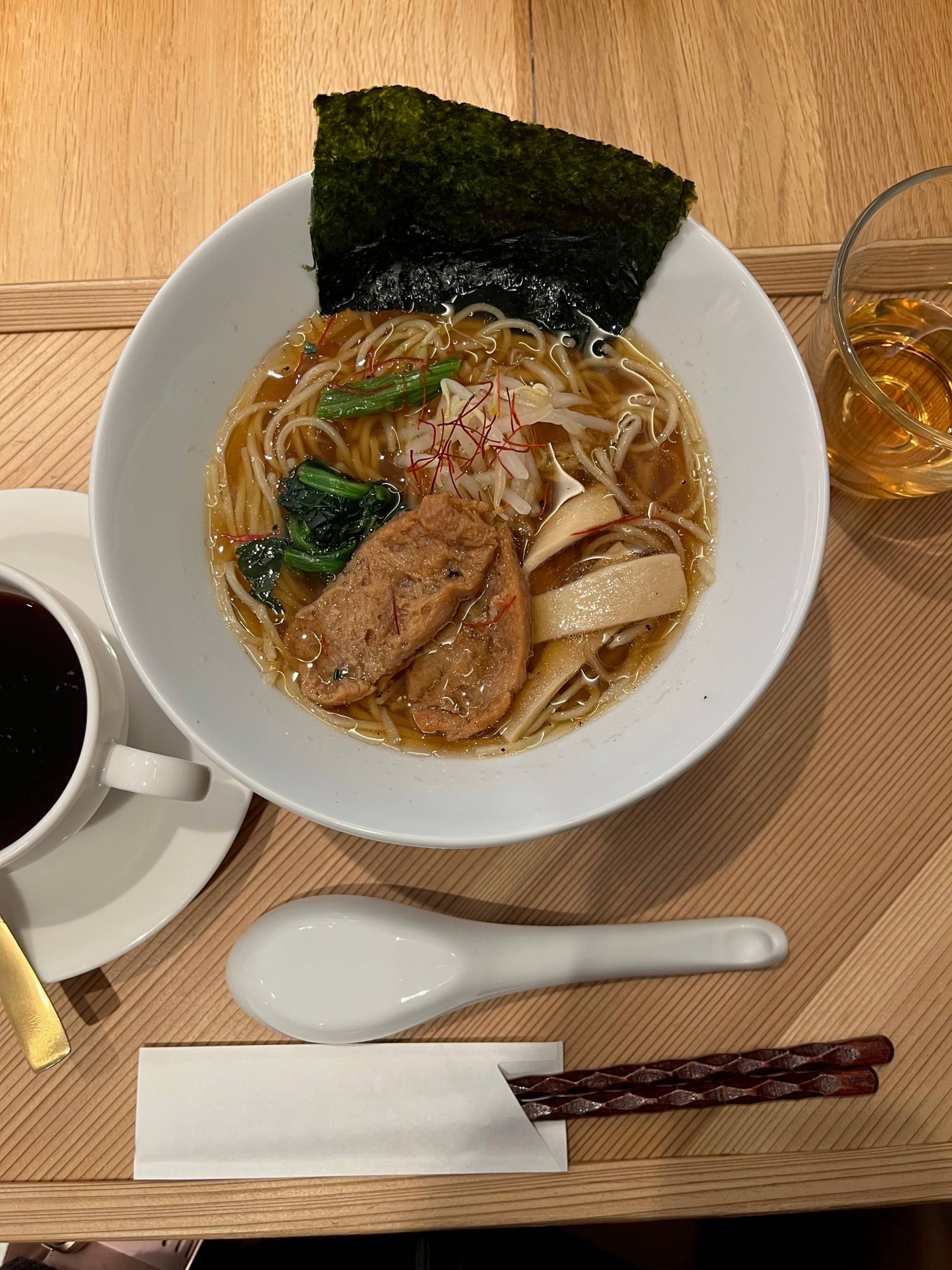 Biała miska japońskiej zupy ramen z makaronem, warzywami i płatem nori stoi na drewnianym stole obok łyżki, pałeczek i napoju.