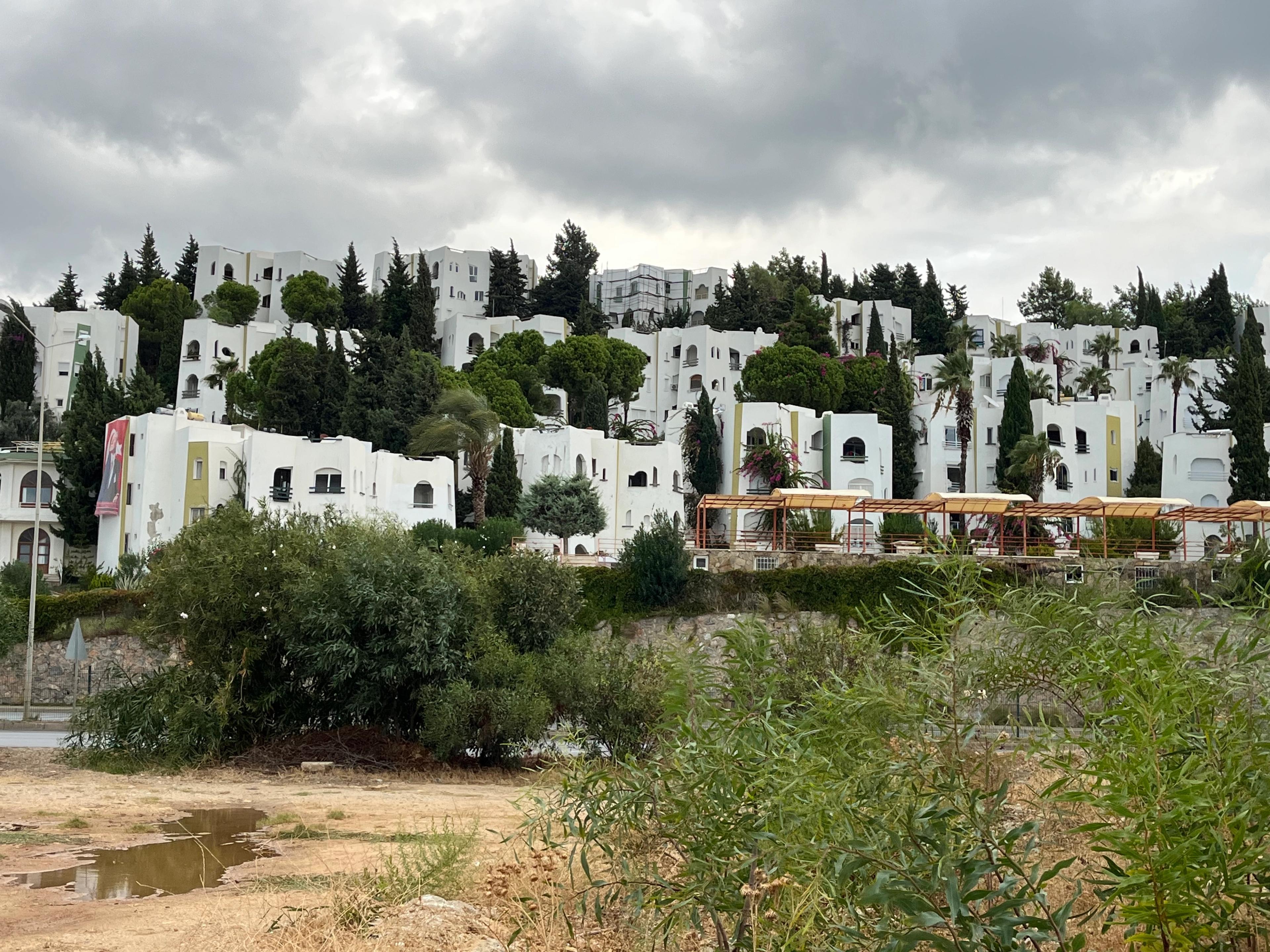 Widok na wzgórze zabudowane białymi budynkami w stylu śródziemnomorskim, otoczonymi zielonymi drzewami i krzewami. Na pierwszym planie widać fragment suchego terenu z roślinnością i kałużą, a w tle gęste, ciemne chmury zasłaniają niebo. Budynki mają łukowe okna i drzwi, a pomiędzy nimi rosną wysokie cyprysy.