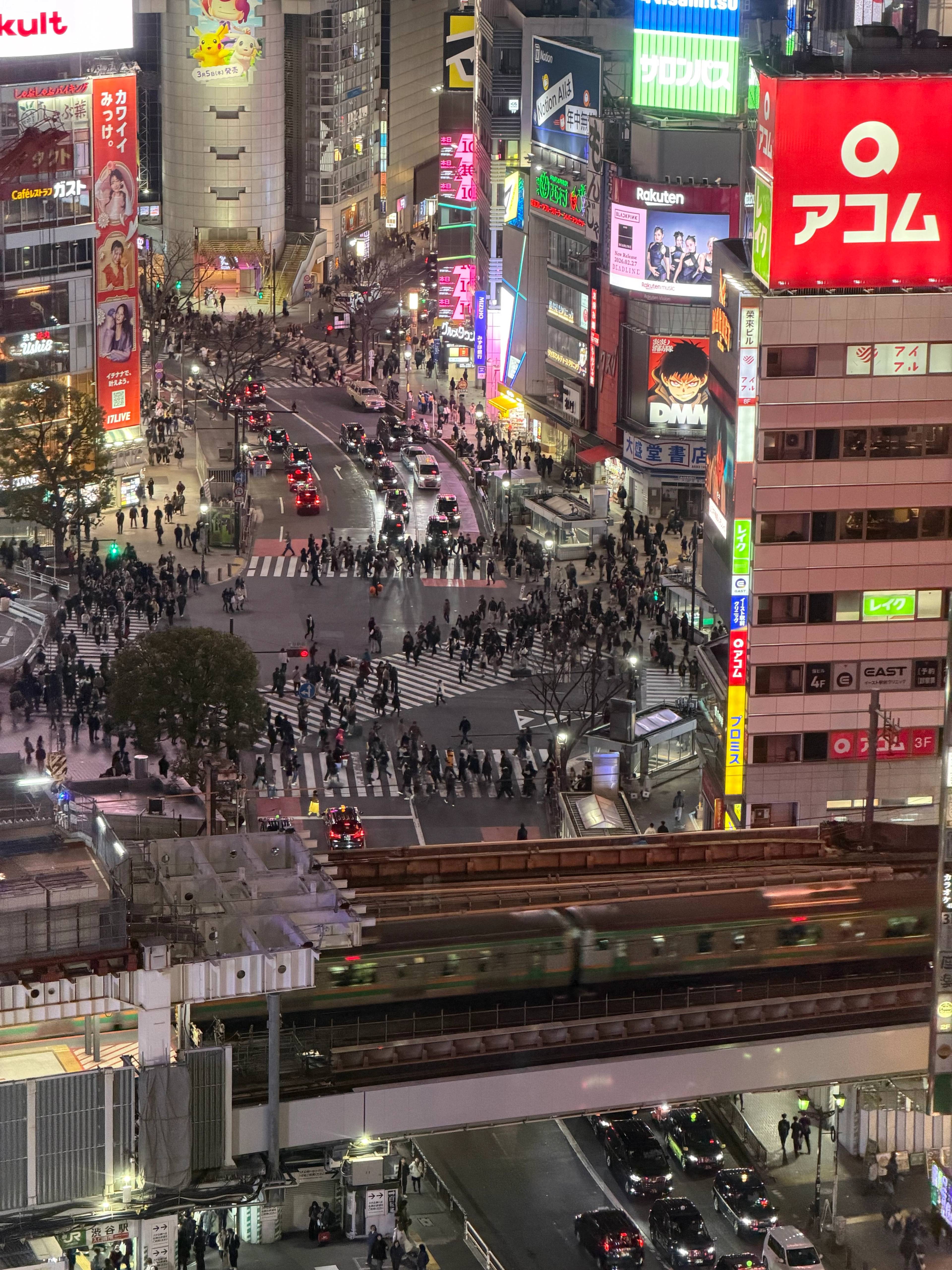 Widok na Shibuya Crossing w Tokio nocą z tłumem pieszych, neonowymi reklamami i przejeżdżającym pociągiem na pierwszym planie.