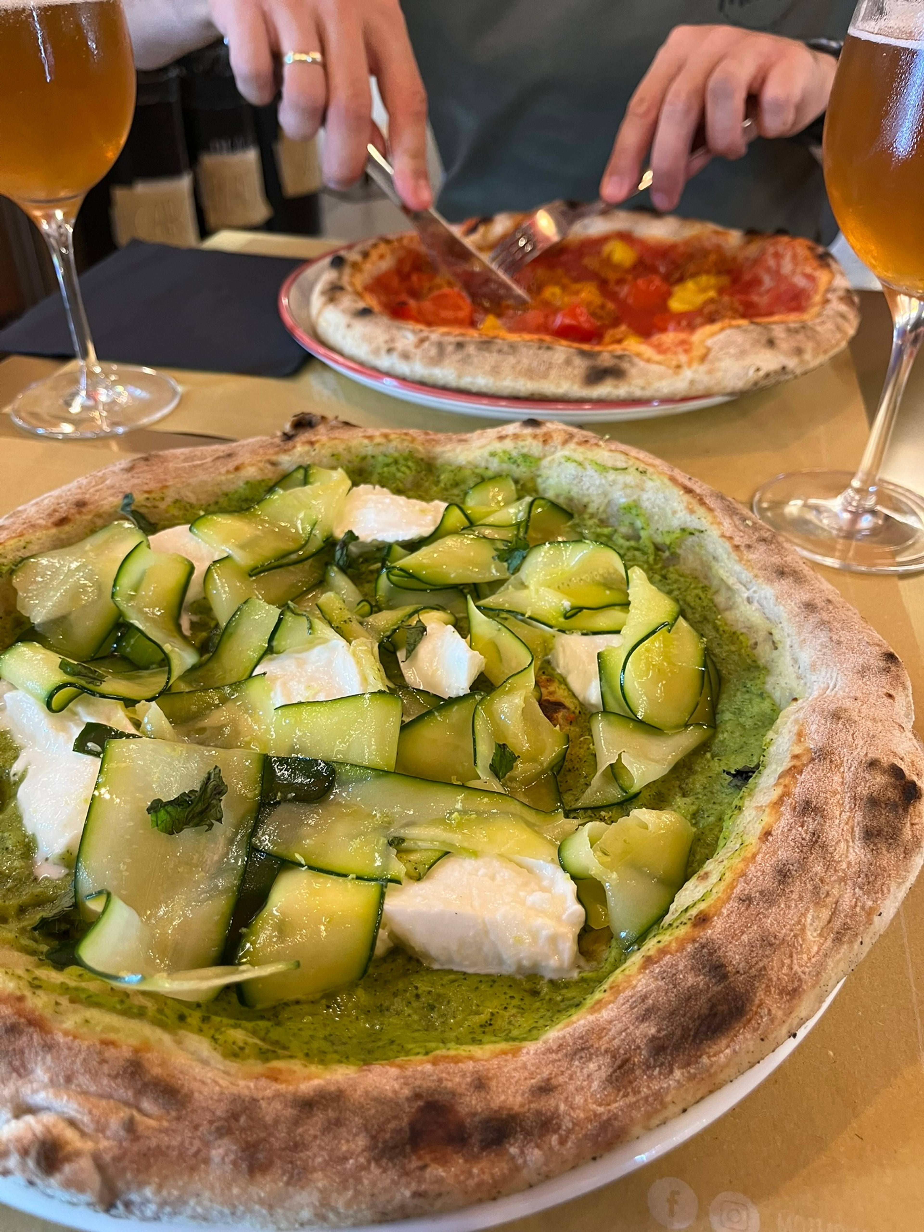 Dwie pizze na puchatym cieście, na pierwszym planie pizza z zielonym sosem, cukinią i białym serem, na drugim planie czerwona pizza z pomidorami