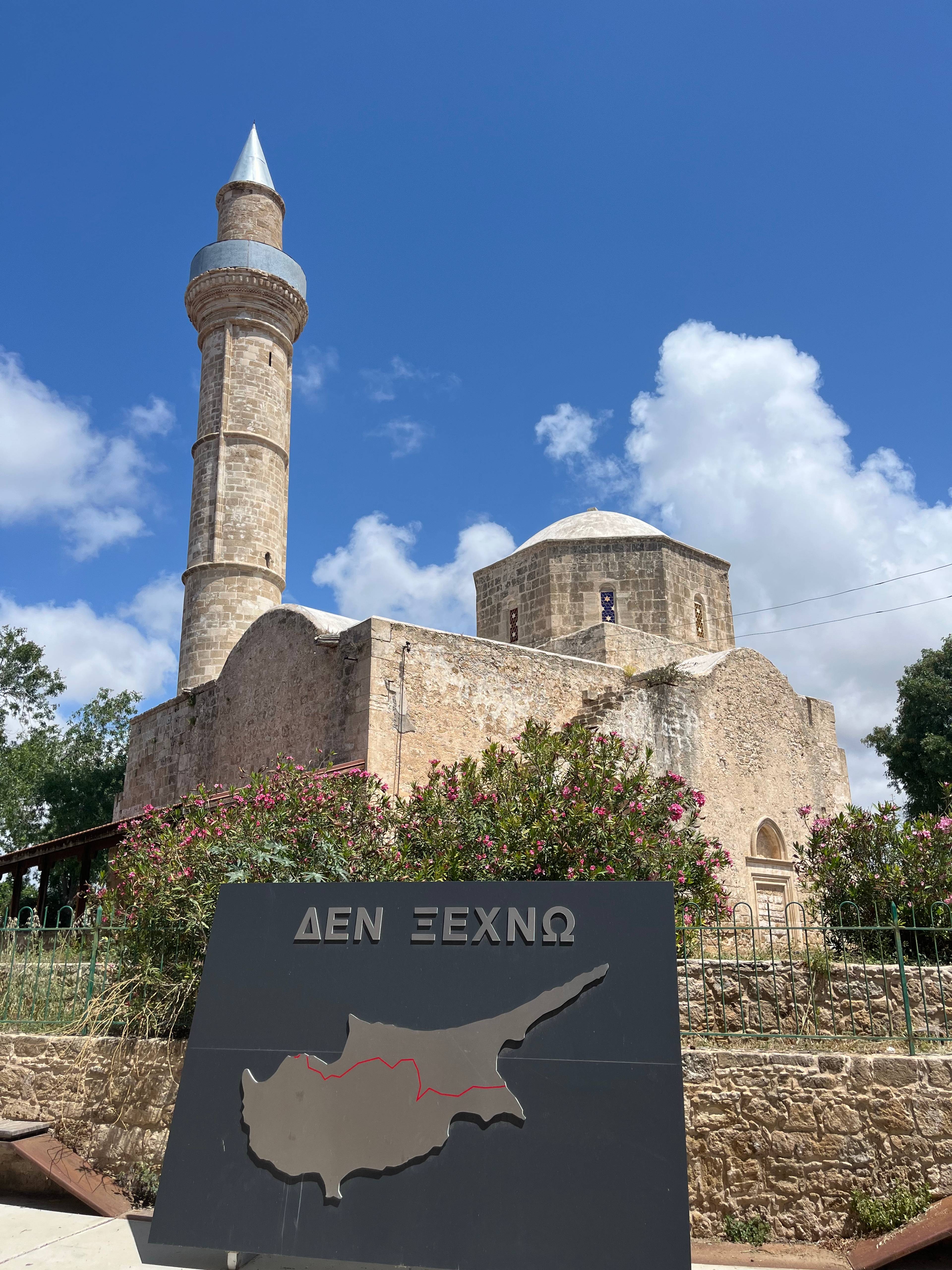 Zabytkowy meczet z wysokim minaretem i kopułą na tle błękitnego nieba z chmurami; na pierwszym planie znak z konturem Cypru, czerwoną linią i greckim napisem 'ΔΕΝ ΞΕΧΝΩ'.