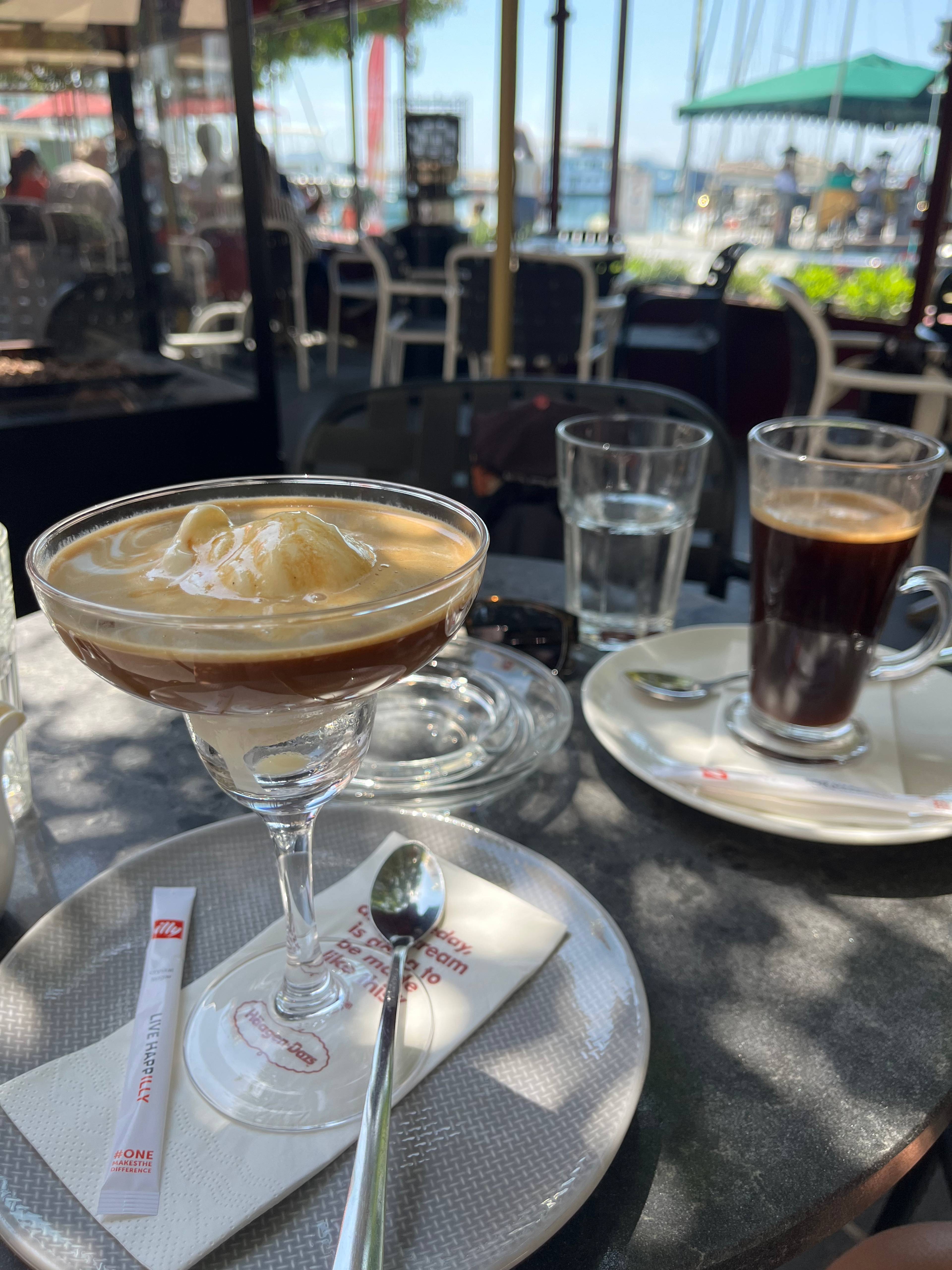 Stolik w kawiarni z affogato w kieliszku, czarną kawą w wysokiej szklance, wodą i widokiem na marinę w tle.