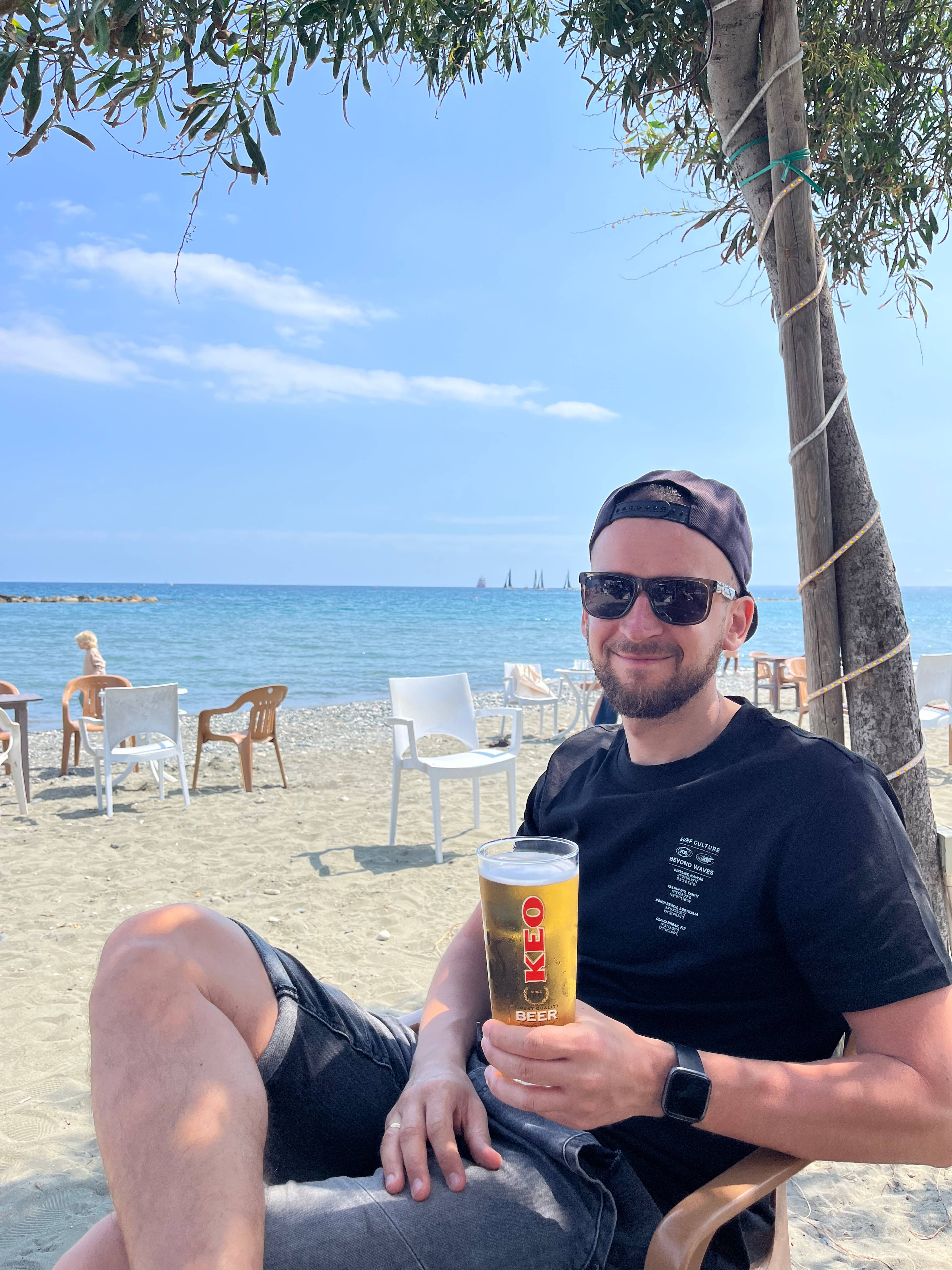 Osoba siedząca na plaży pod drzewem z piwem w ręku, w tle morze, niebo i stoliki z ludźmi.