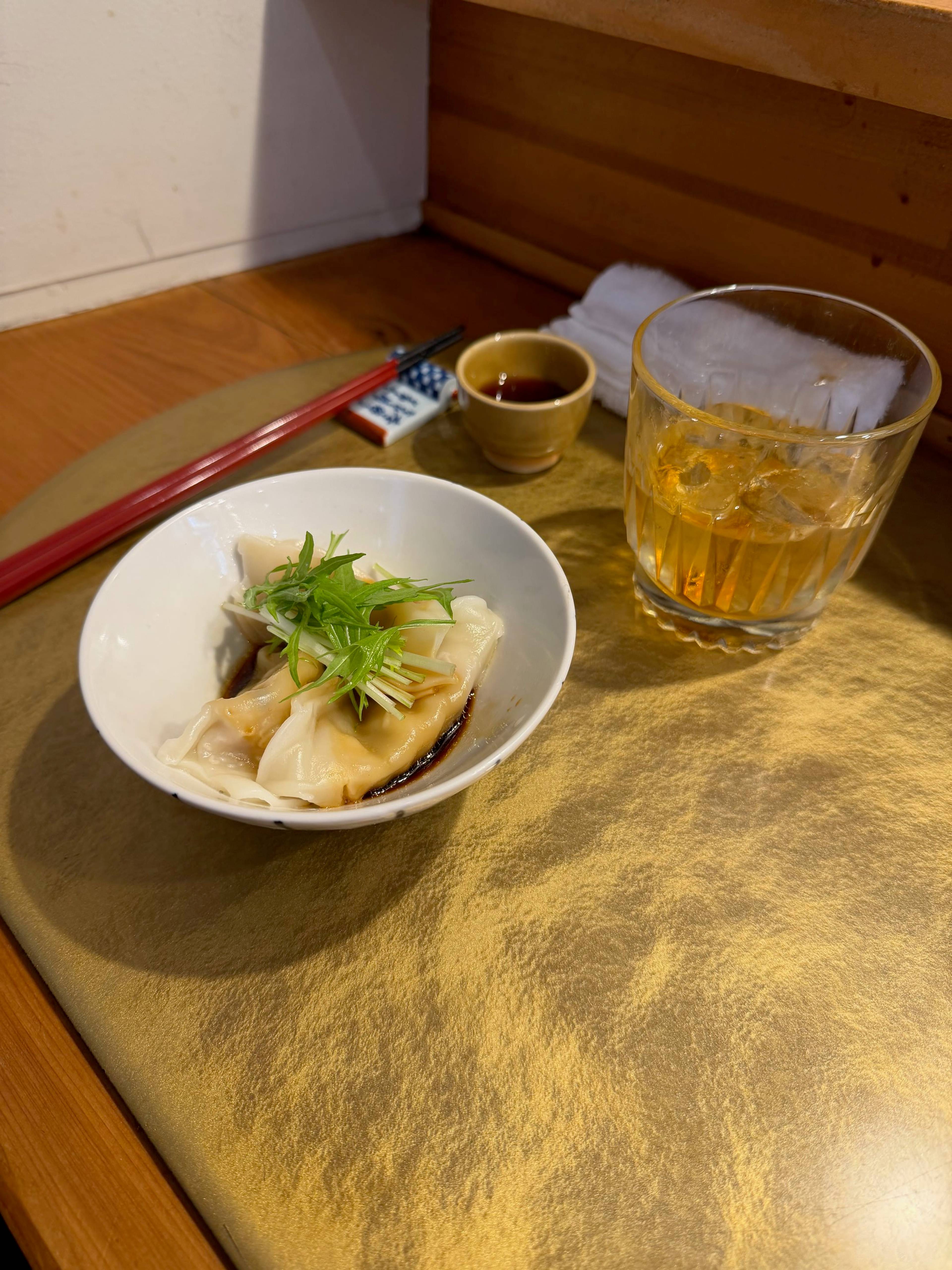 Niewielka miseczka z japońskim pierożkiem gyoza stoi na złotej tacce obok szklanki sake, pałeczek i małej miseczki z sosem sojowym.