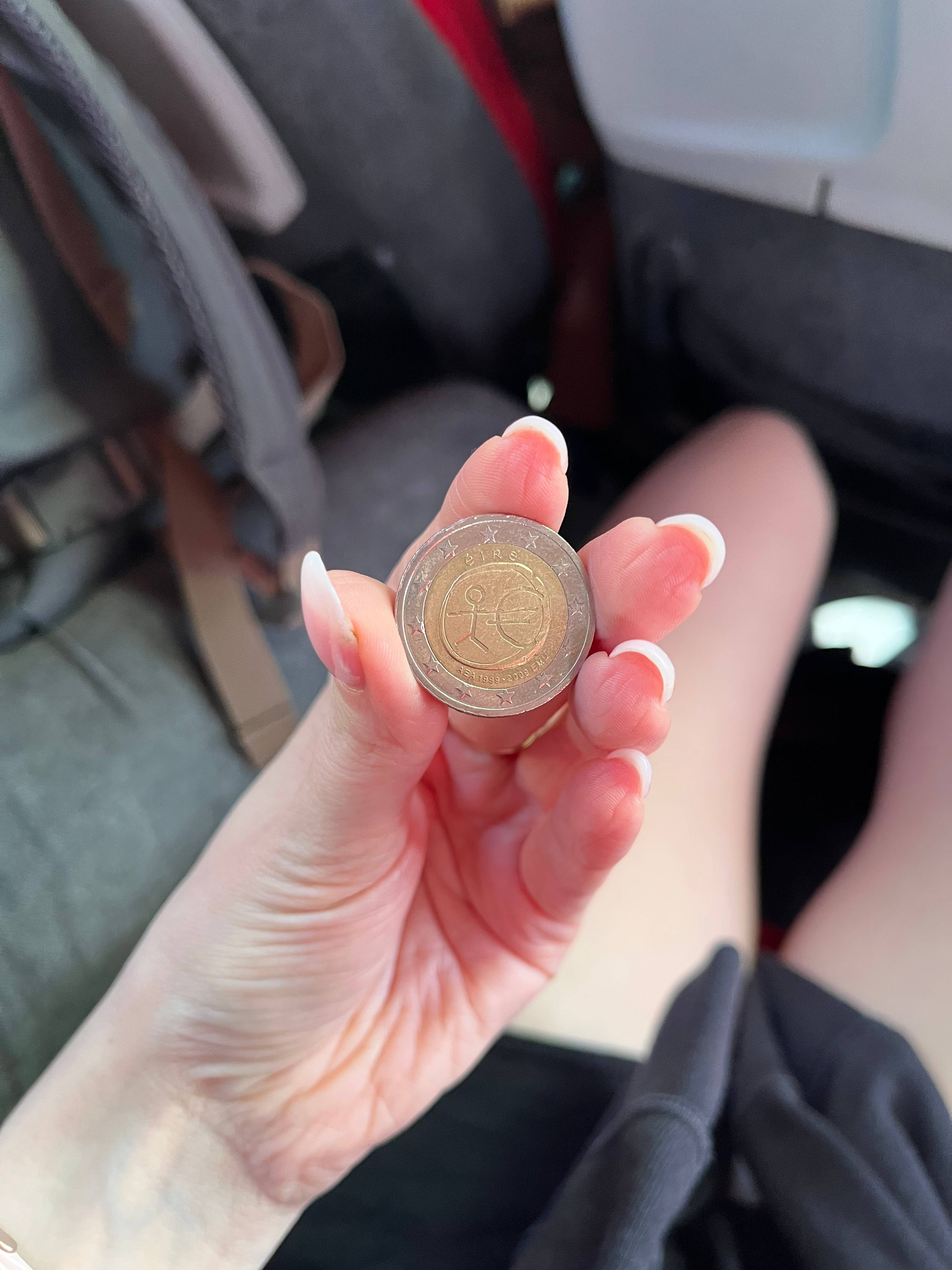 Zbliżenie na dłoń trzymającą dwukolorową monetę z widocznym wzorem w centrum. W tle widać fragment siedzenia i plecaka, co sugeruje, że zdjęcie zostało wykonane w pojeździe. Oświetlenie jest naturalne, a ostrość skupiona na monecie i dłoni.