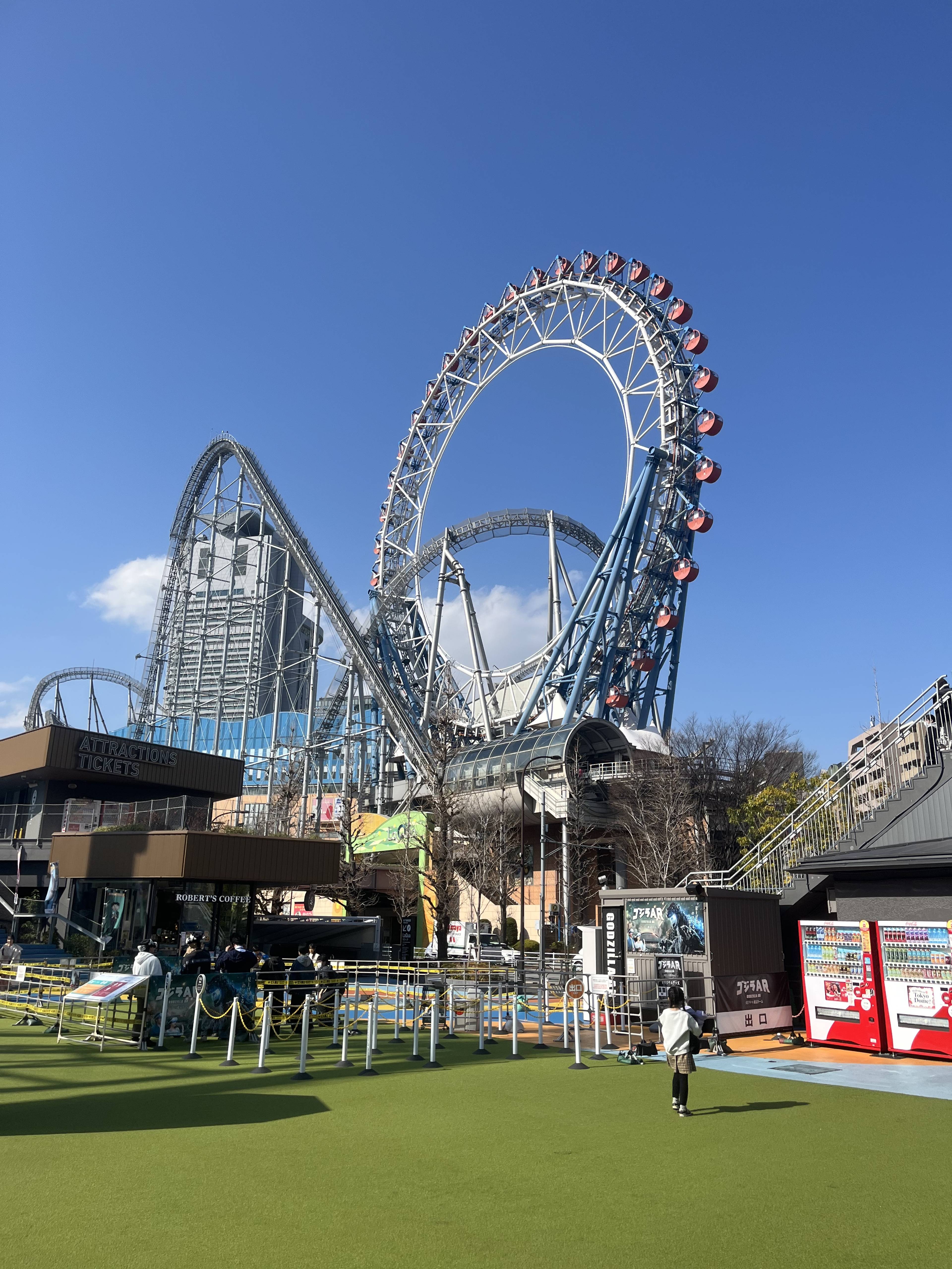 Duża stalowa kolejka górska z pętlą oraz diabelski młyn widoczne na terenie parku rozrywki Tokyo Dome pod bezchmurnym niebem.