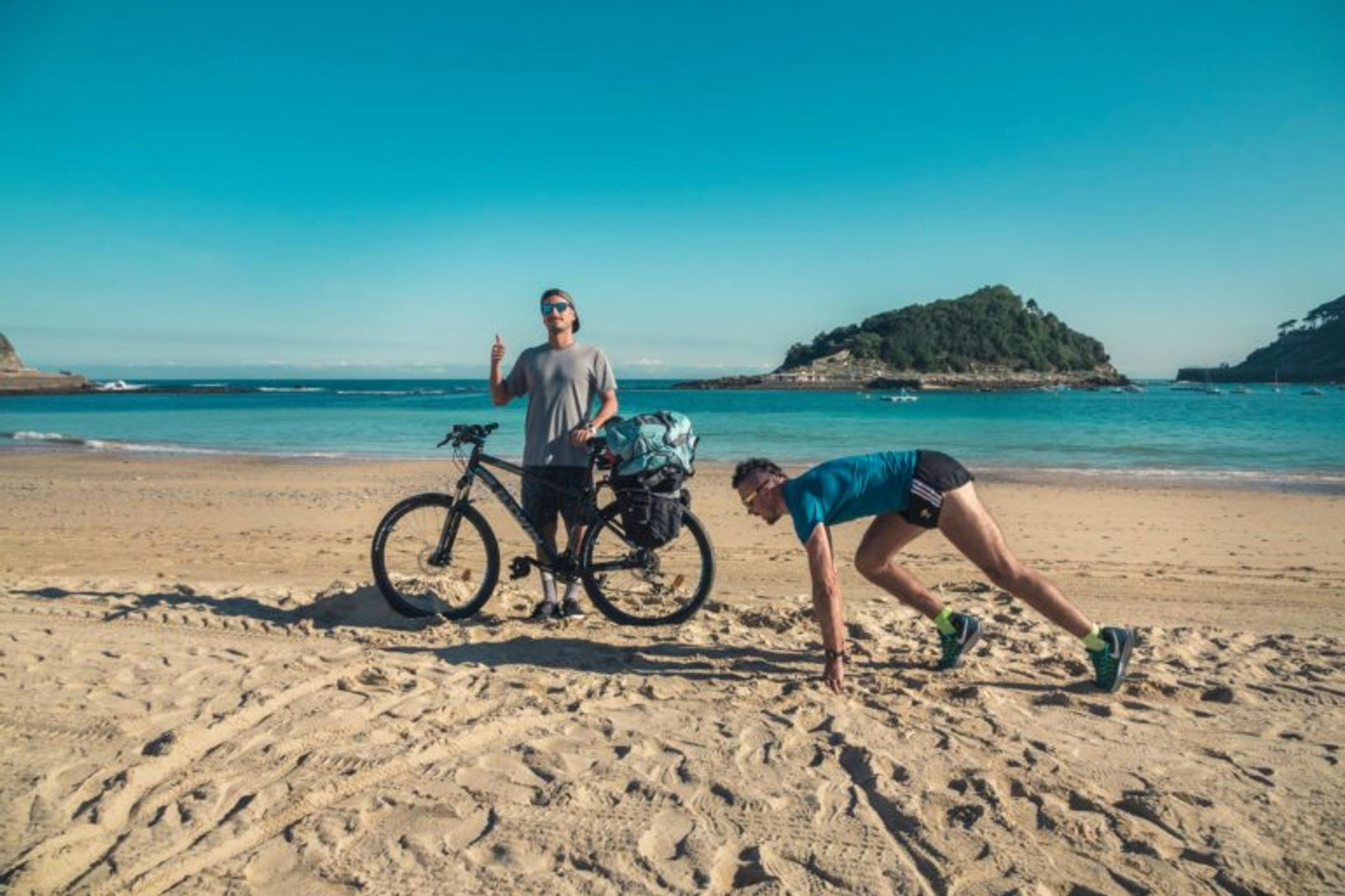 Na plaży stoi dwóch mężczyzn, jeden przy rowerze, drugi w pozycji jak do startu biegu. W tle woda i wyspa. 
