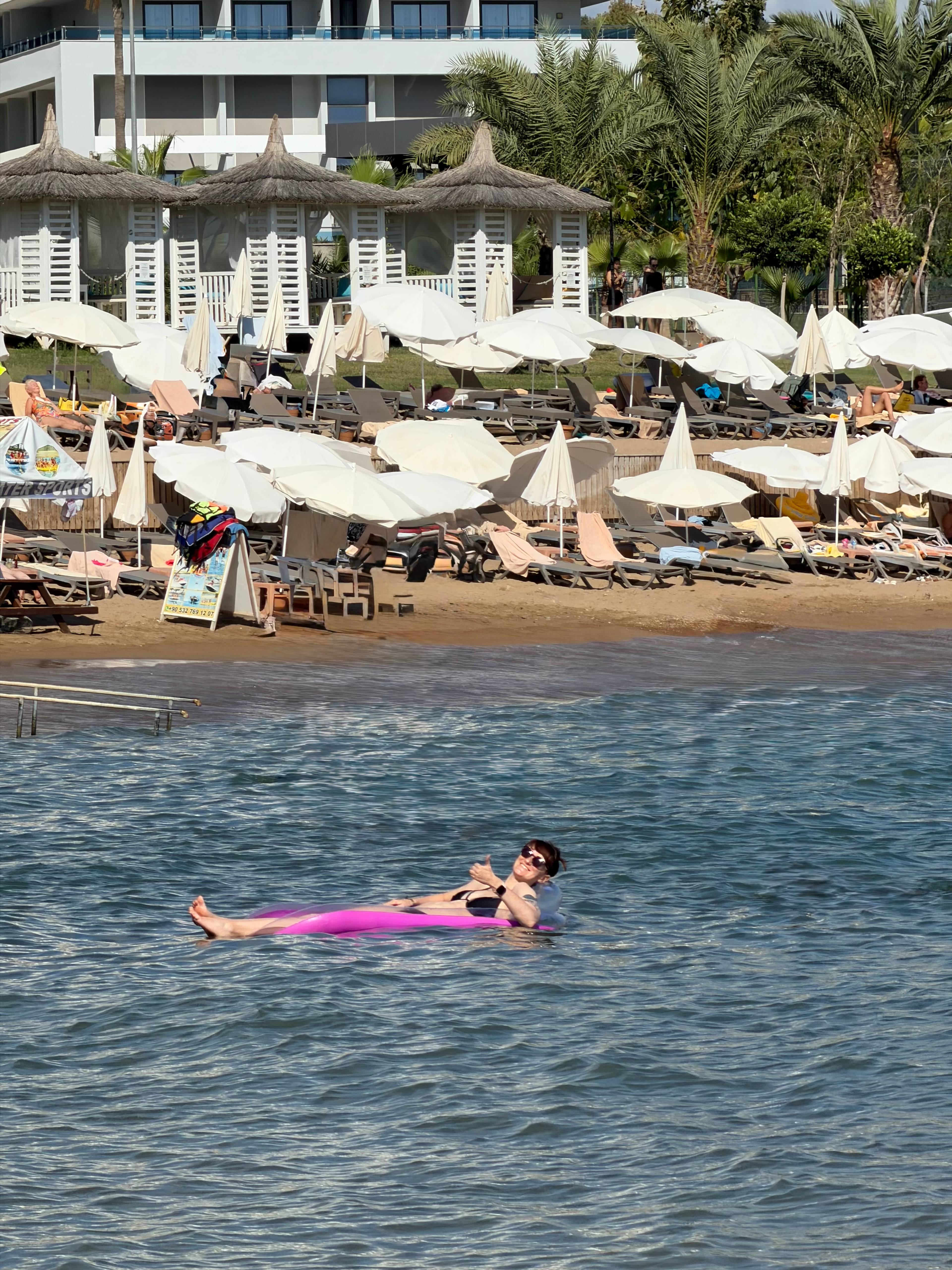 Osoba na dmuchanym materacu w wodzie blisko plaży z leżakami i parasolami w tle.