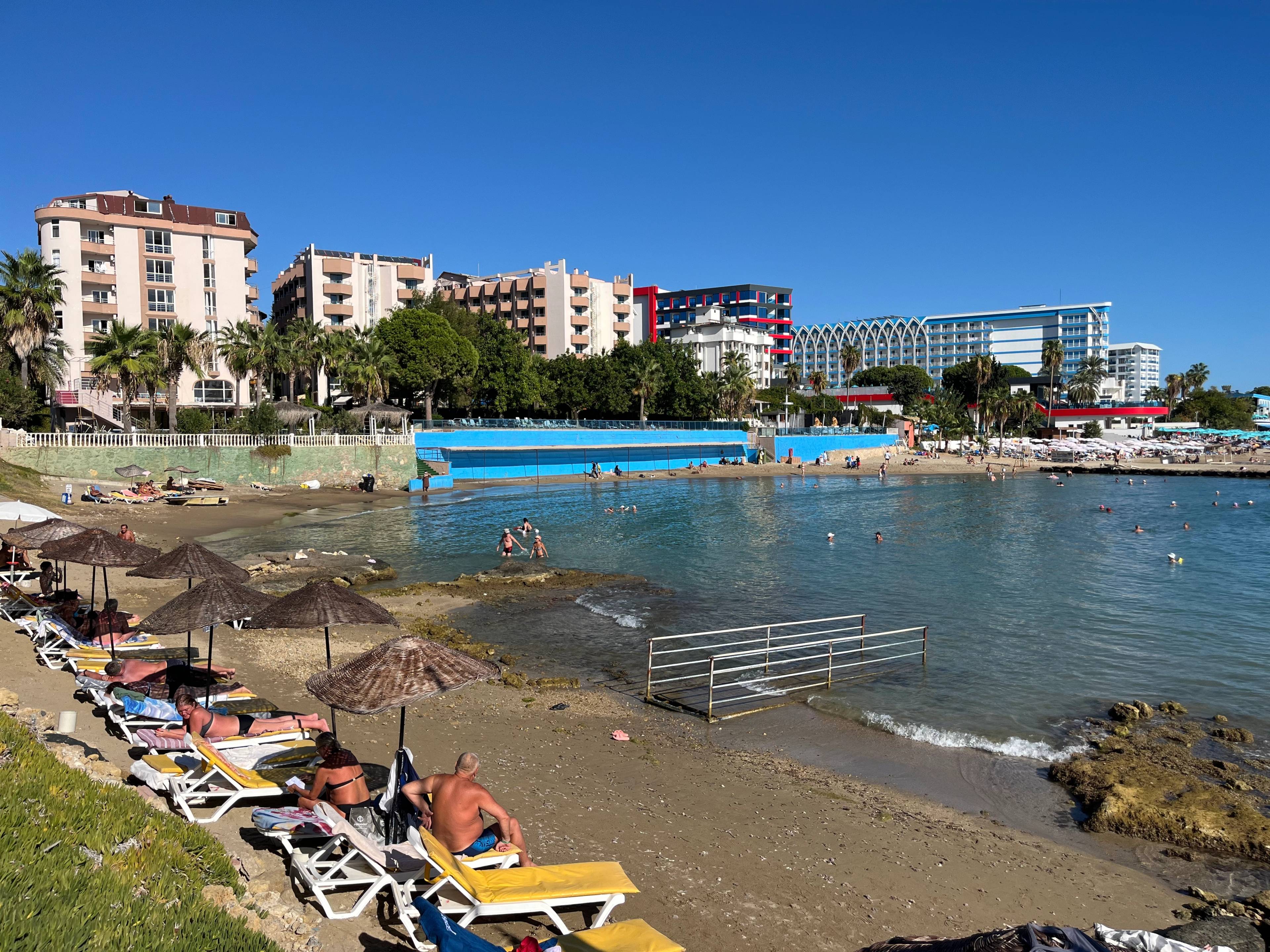 Plaża z leżakami i parasolami nad spokojną zatoką, w tle nowoczesne hotele i apartamentowce pod bezchmurnym, niebieskim niebem.