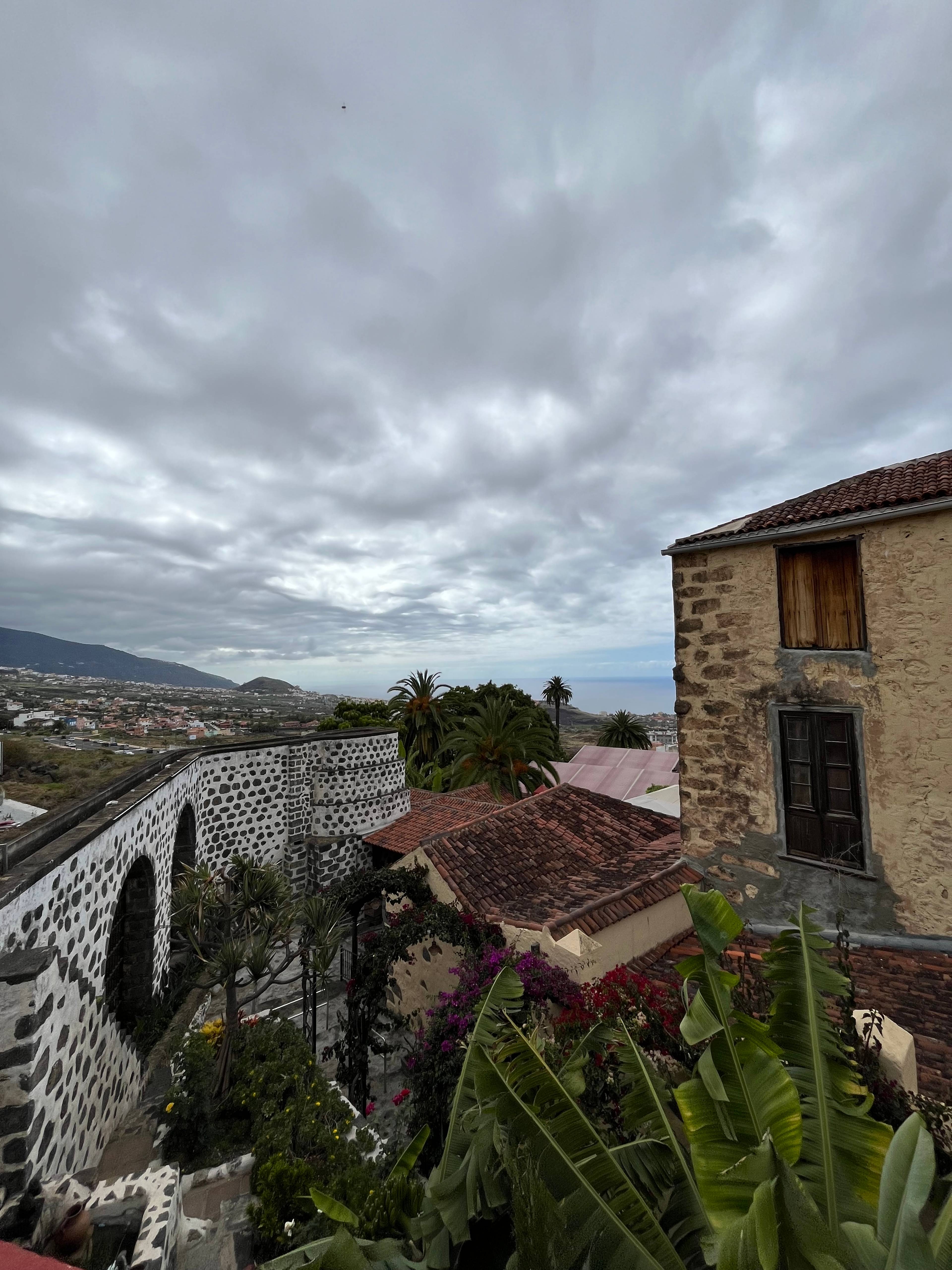 Kamienne budynki i fragment młyna na tle pochmurnego nieba i zielonej roślinności.