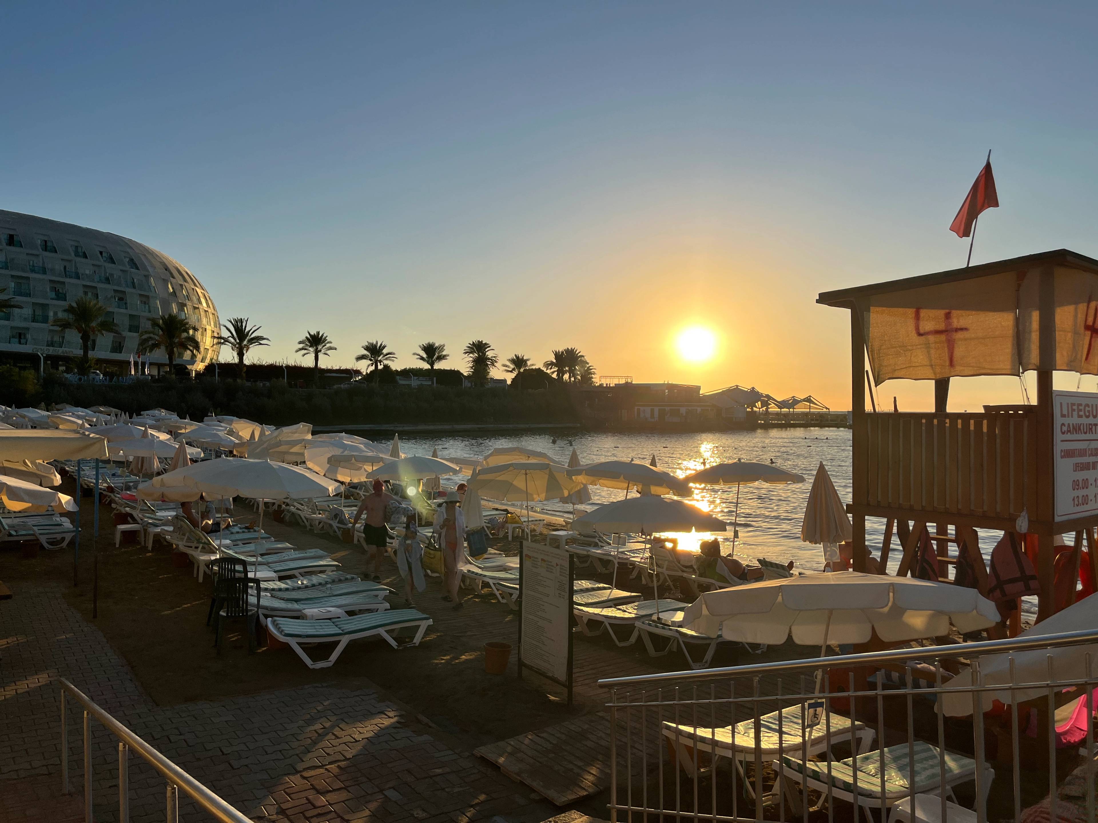 Plaża z rzędami leżaków i parasoli, drewniana wieża ratownika z czerwoną flagą, a w tle morze i zachodzące słońce nad horyzontem.