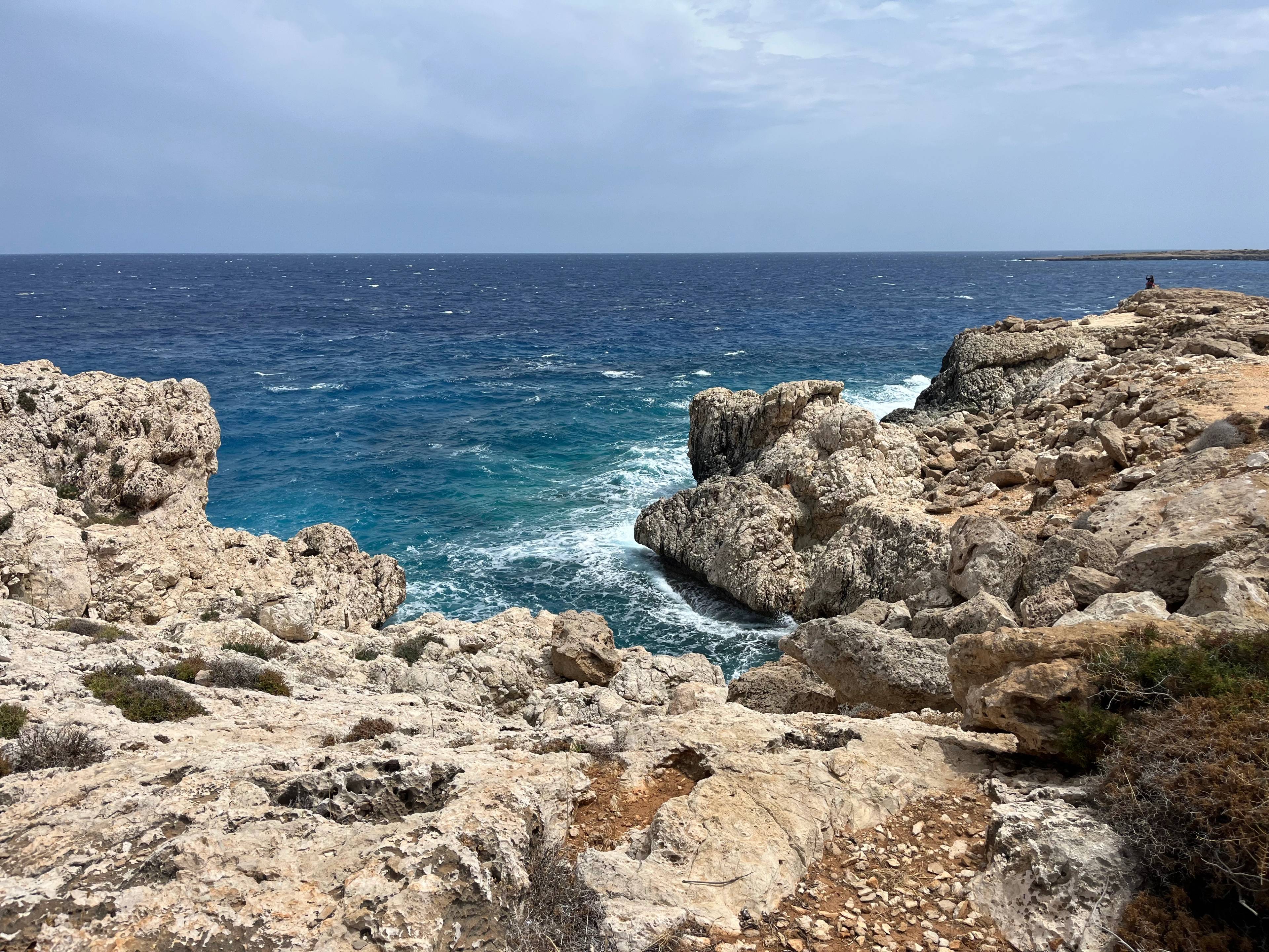 Skaliste wybrzeże Capo Greco na Cyprze z turkusowym morzem i falami uderzającymi o skały.