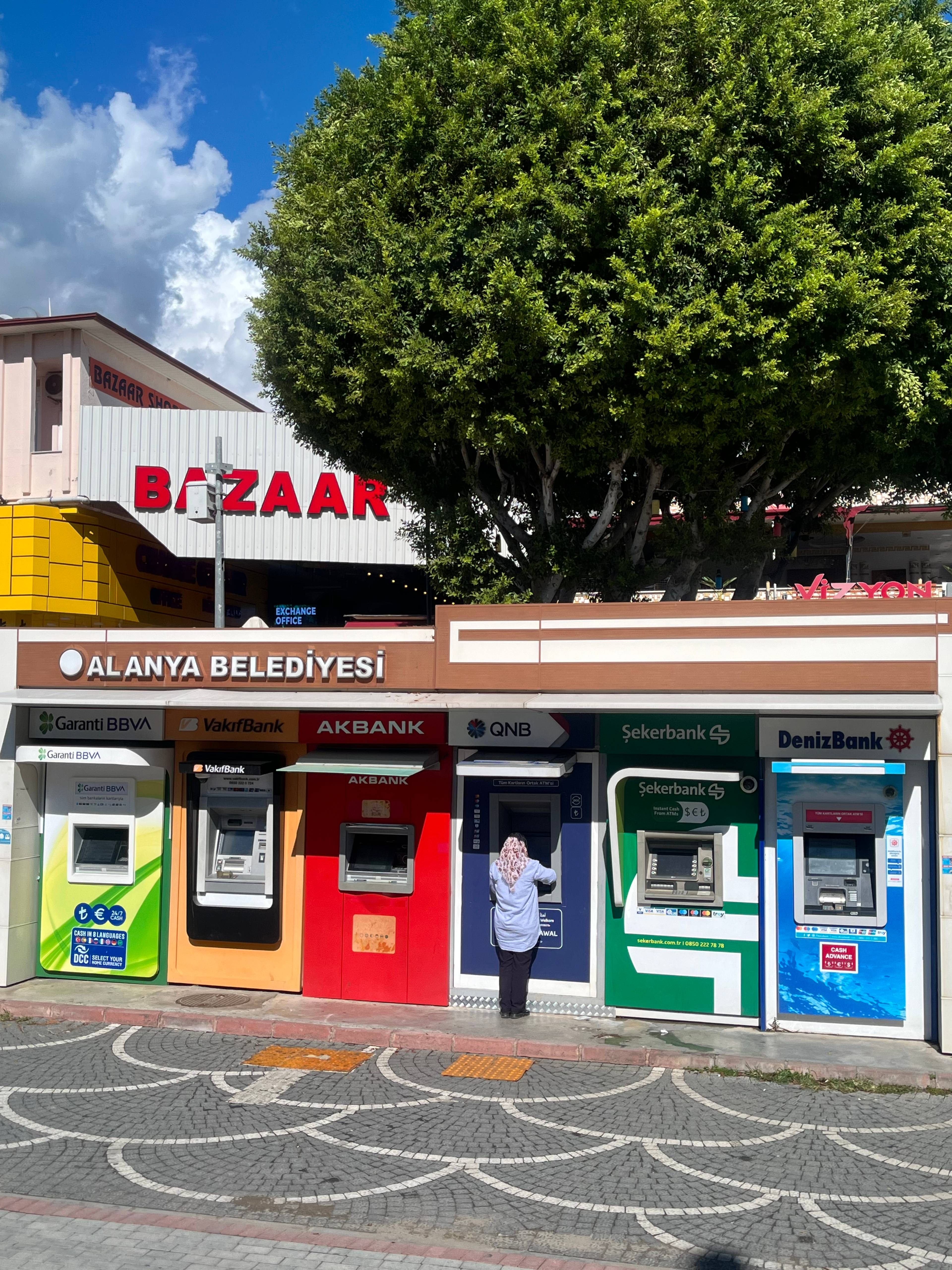 Kolorowe bankomaty ustawione w rzędzie na chodniku przy ulicy. w Tle blaszany budynek z napisem "Bazaar"